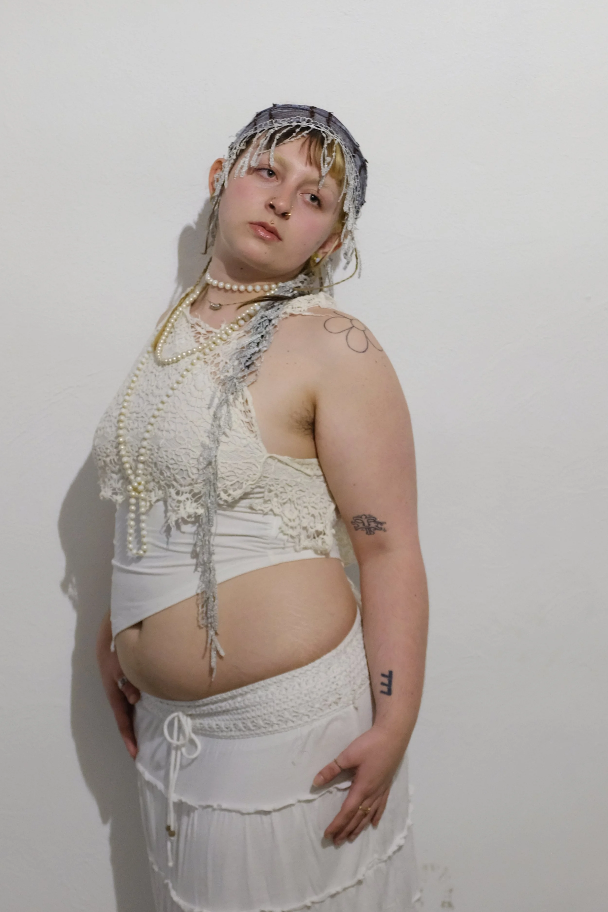 A young woman with tattoos and piercings posing against a plain wall, wearing a lace top, a skirt, and layered pearl necklaces, with a headscarf and short hair.