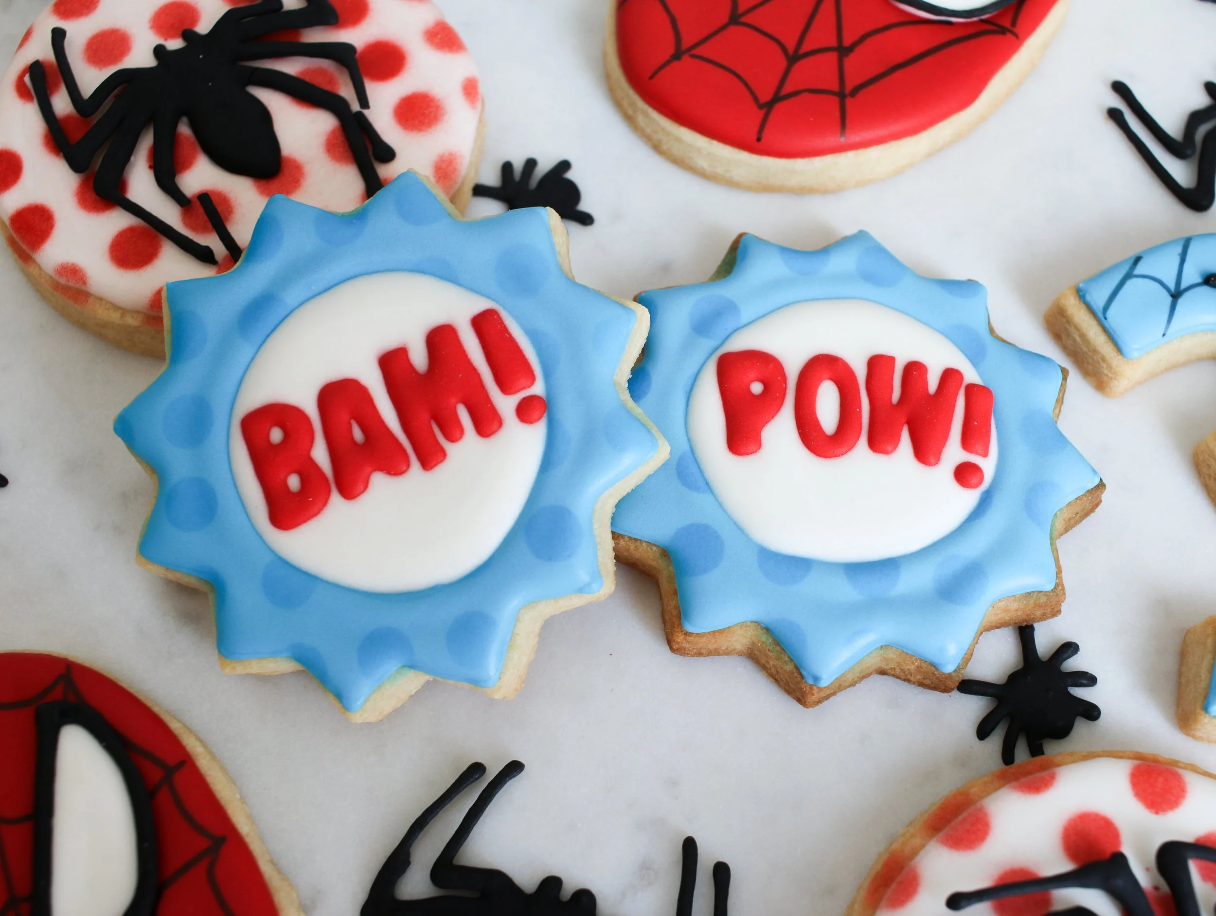 Decorated cookies with comic book style words 'BAM!' and 'POW!' in red icing on white backgrounds, with blue and red themed spider web and spider designs.