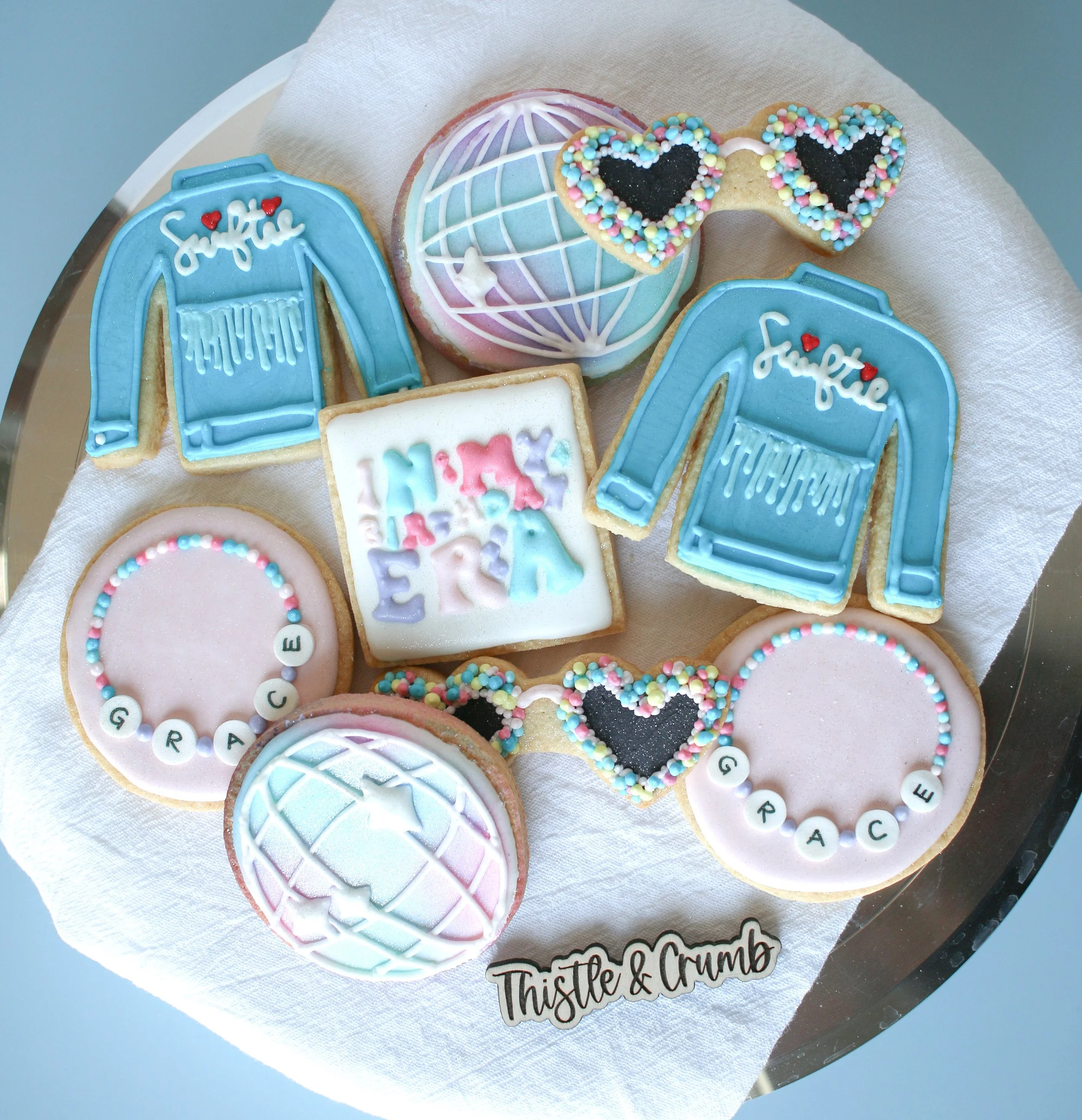 Assorted decorated cookies on a tray, including denim jackets, globes, friendship bracelets, heart sunglasses with sprinkles, and a square cookie with colorful text, all on a white towel with 'Thistle & Crumb' branding.