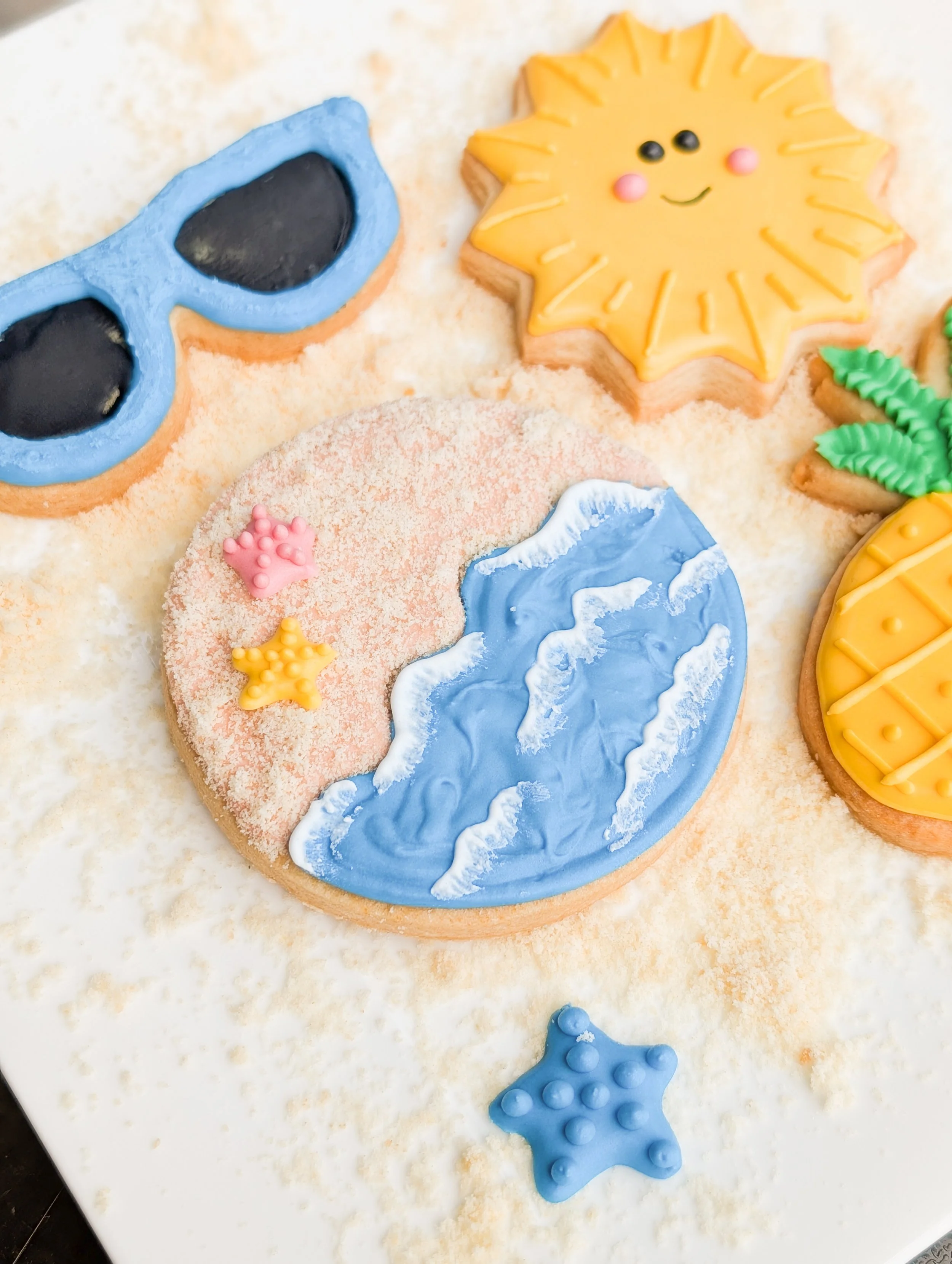 Decorated cookies with summer and beach themes including sunglasses, a sun, a wave, a pineapple, and stars, all on a light surface.