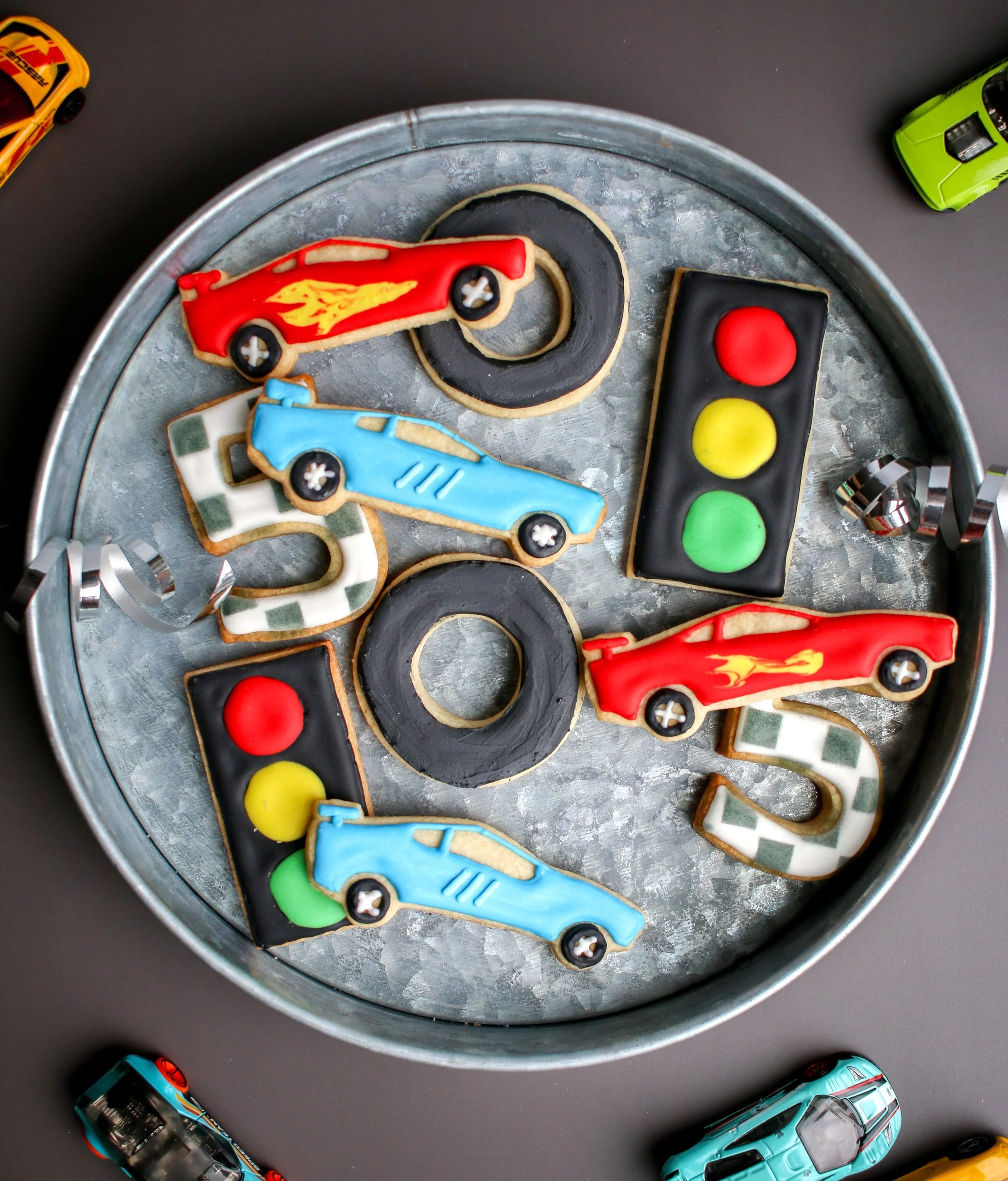 Cookies shaped and decorated as race cars, a traffic light, and the number five, displayed on a round metallic tray, with toy cars around it on a gray surface.