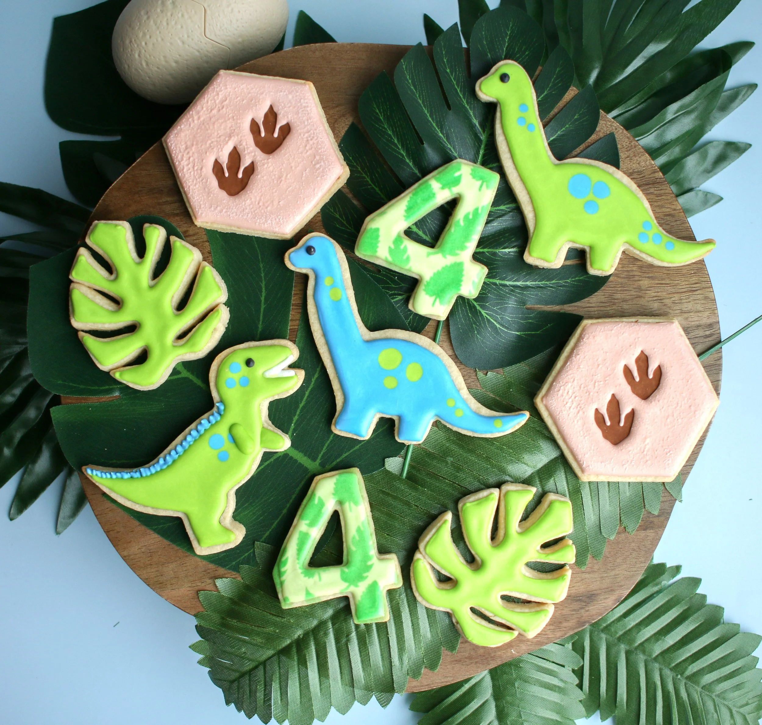 Decorative cookies on wooden tray with green leaves, featuring dinosaur shapes, leafy patterns, and numbers 4 and 5, with pink hexagon-shaped cookies with handprint cutouts.