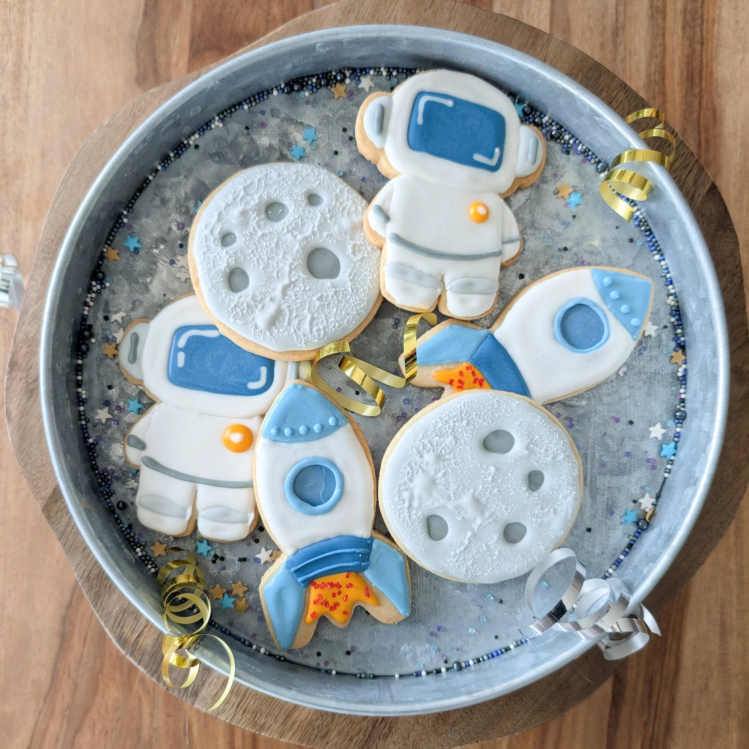Space-themed decorated cookies, including astronauts, rockets, and moons, on a silver tray with star and bead decoration.