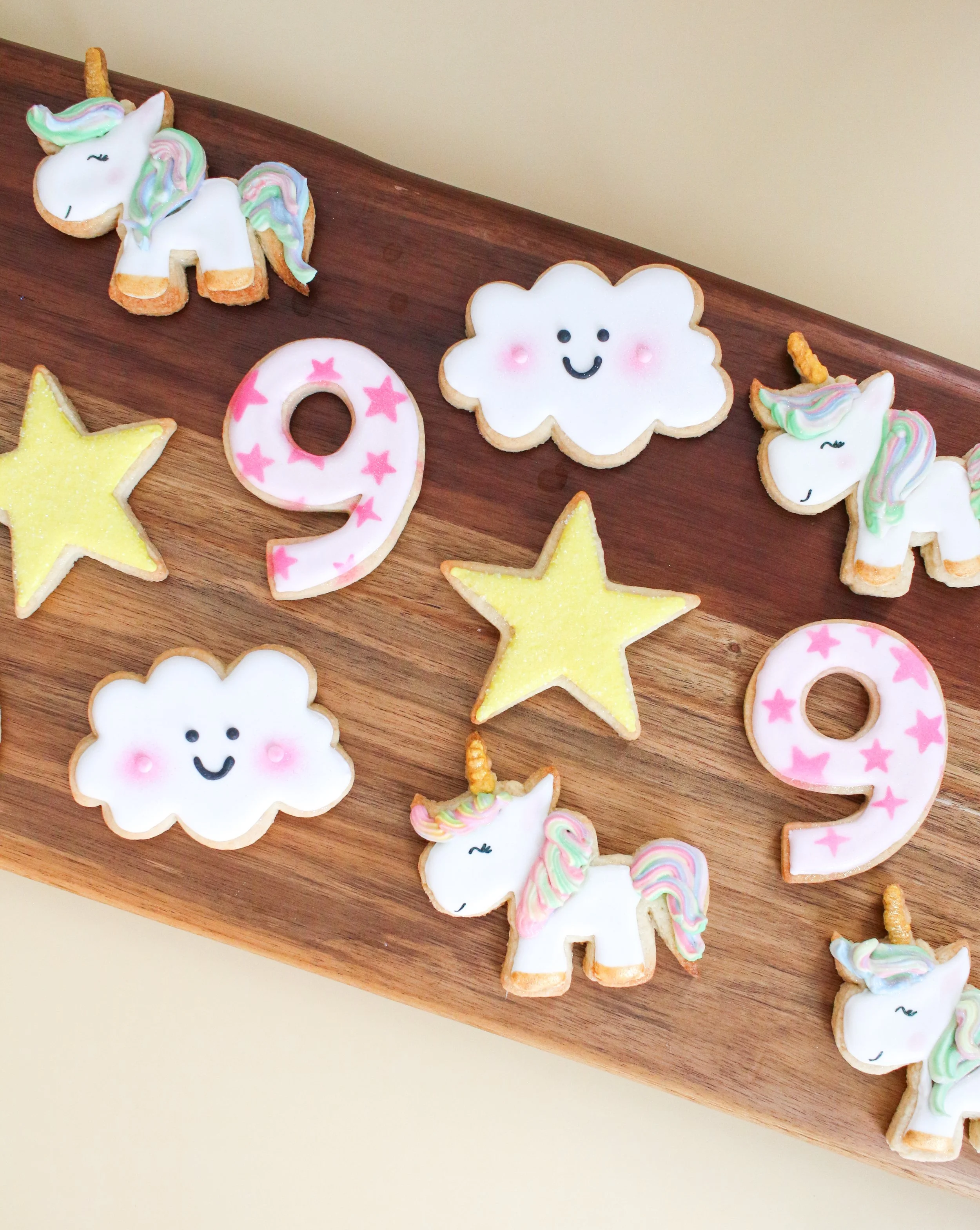 Decorative cookies on a wooden tray, including unicorns, clouds, stars, and the number 9, with pastel-colored icing and cheerful faces.