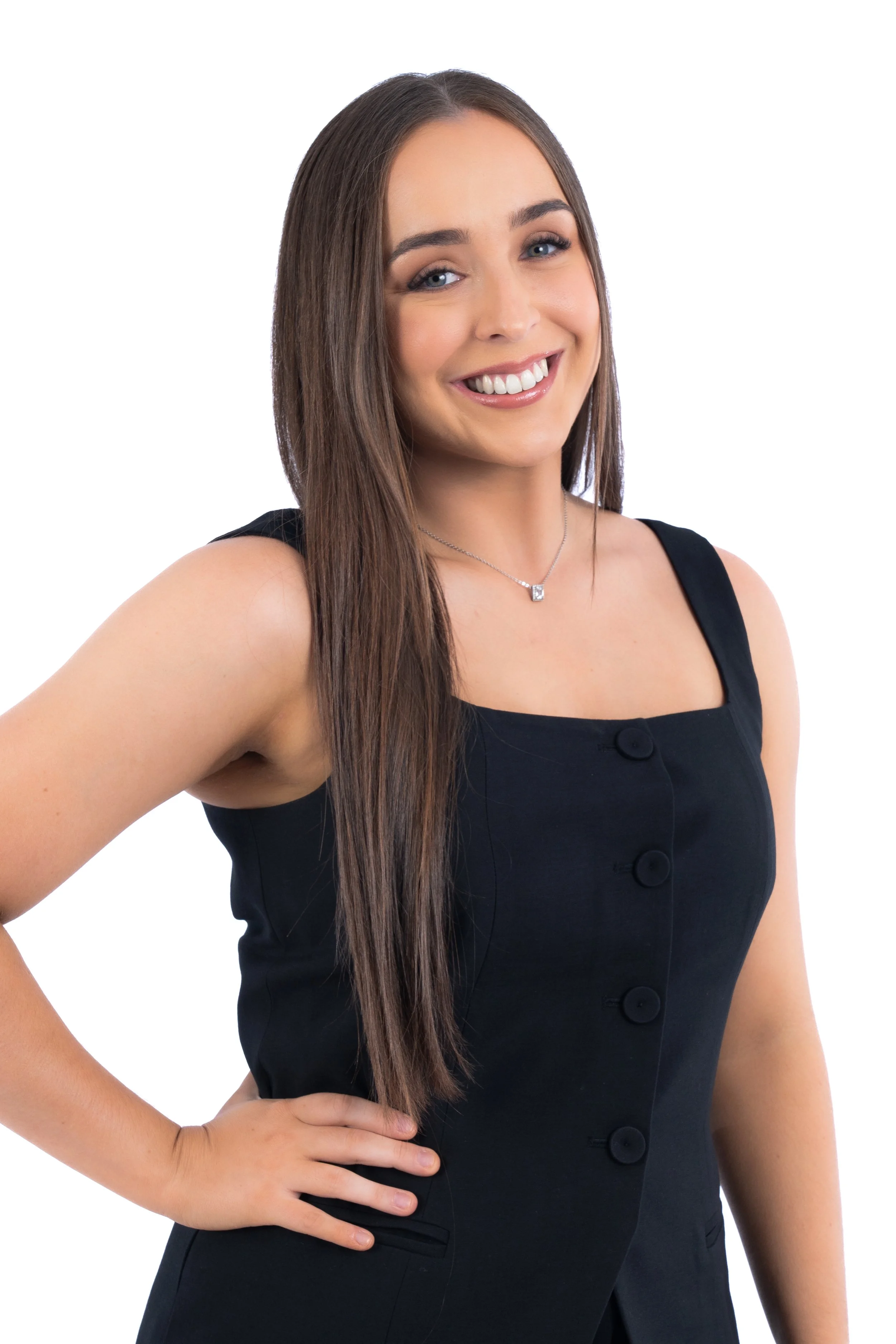 A young woman with long brown hair, smiling, wearing a black sleeveless dress with buttons and a silver necklace, posing with her hand on her hip against a white background.