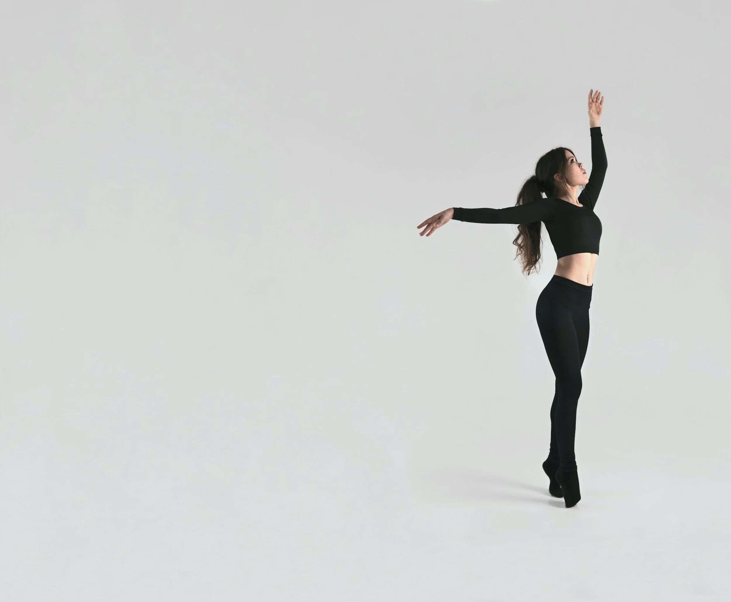 A woman in black athletic wear standing on tiptoe with her arms extended in a dance pose against a plain white background.
