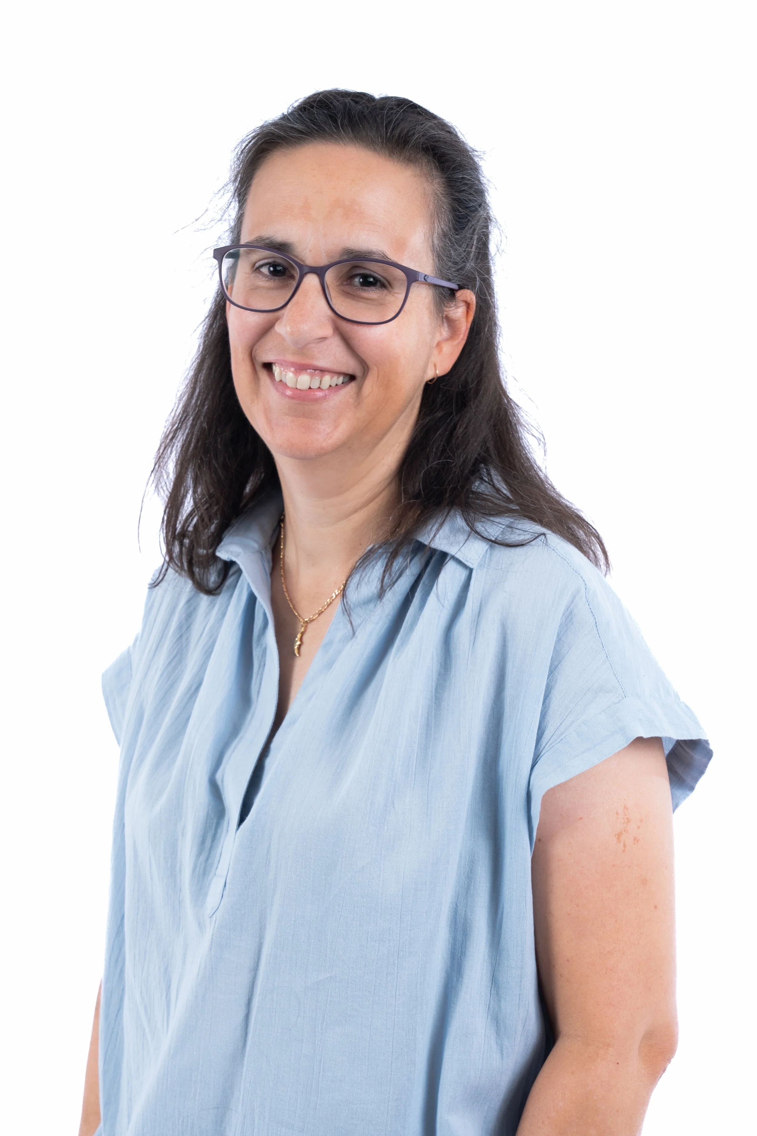 A woman with long dark hair, wearing glasses, a light blue blouse, and a gold necklace, smiling against a white background.