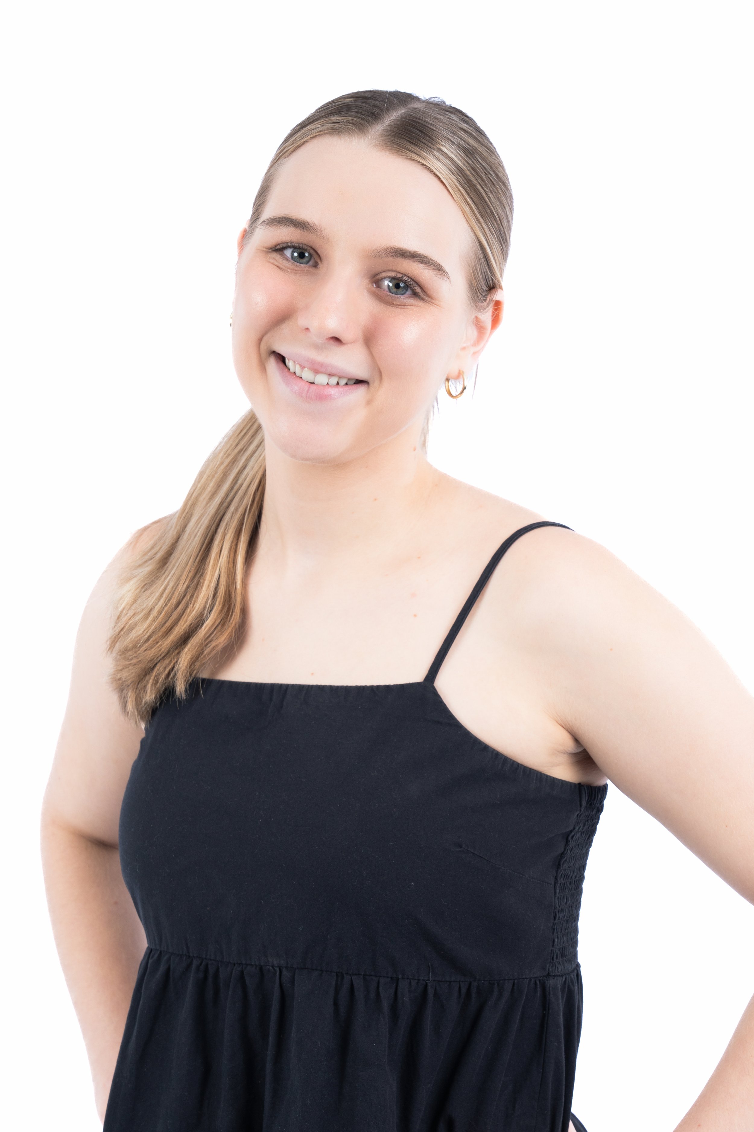 A young woman with long light brown hair in a ponytail, wearing a black sleeveless dress and small hoop earrings, smiling at the camera against a white background.