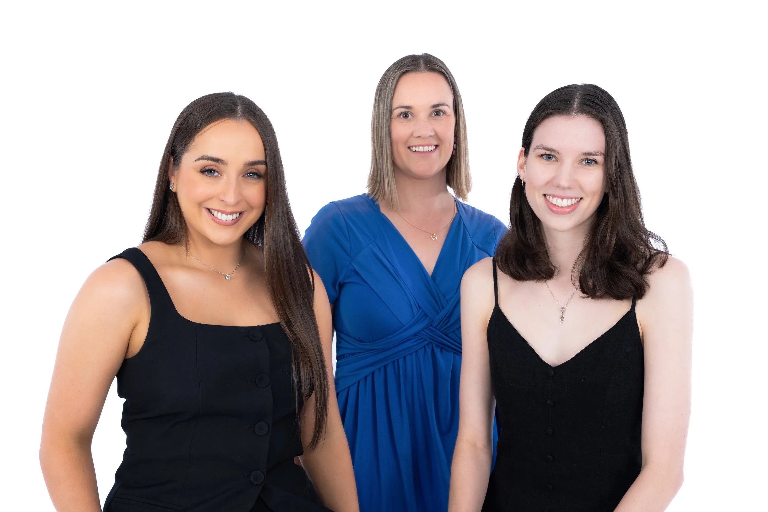 Three women smiling, two with dark brown hair and one with blonde hair, wearing sleeveless dresses blue and black against a white background.