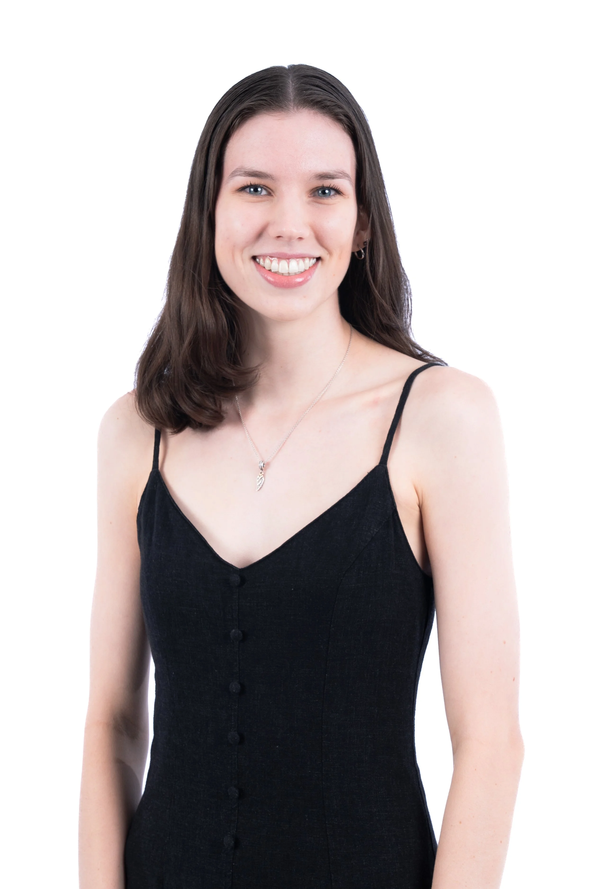 A young woman with shoulder-length dark brown hair, smiling, wearing a black sleeveless dress with buttons and a silver pendant necklace, standing against a plain white background.