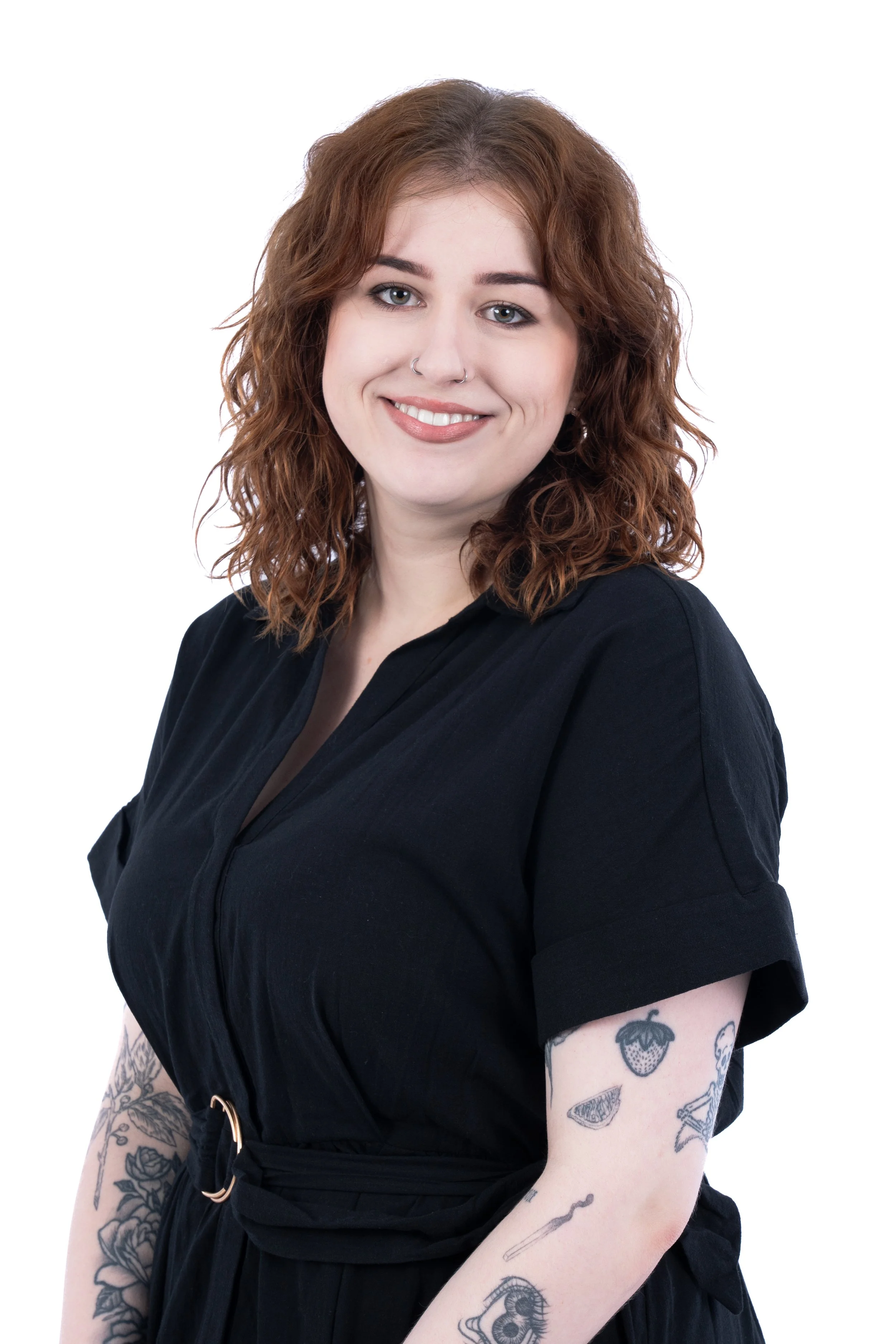 A woman with wavy, shoulder-length red hair, wearing a black dress, smiling with visible tattoos on both arms.