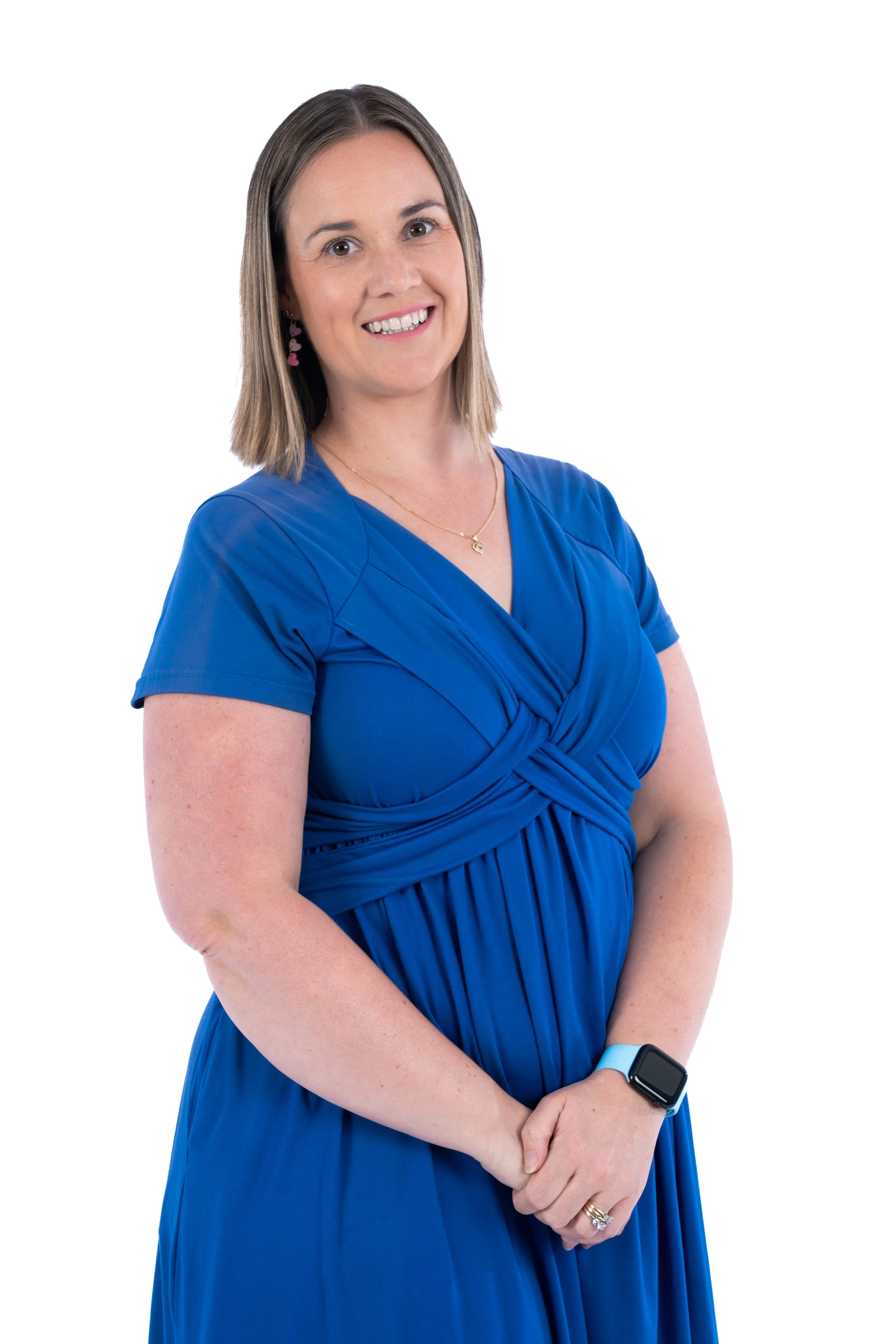 A woman with shoulder-length brown hair, wearing a blue dress, smiling, with a smartwatch on her left wrist and pink earrings, standing against a white background.