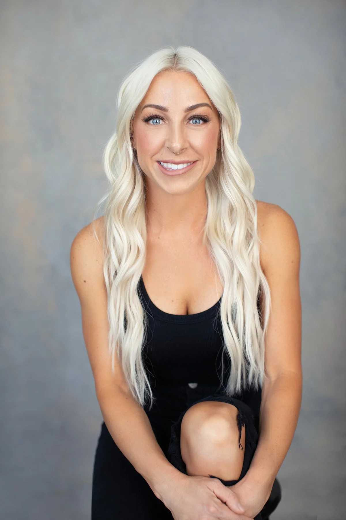 A woman with long blonde wavy hair, blue eyes, and a nose piercing, smiling and wearing a black tank top and black ripped jeans, sitting against a gray background.