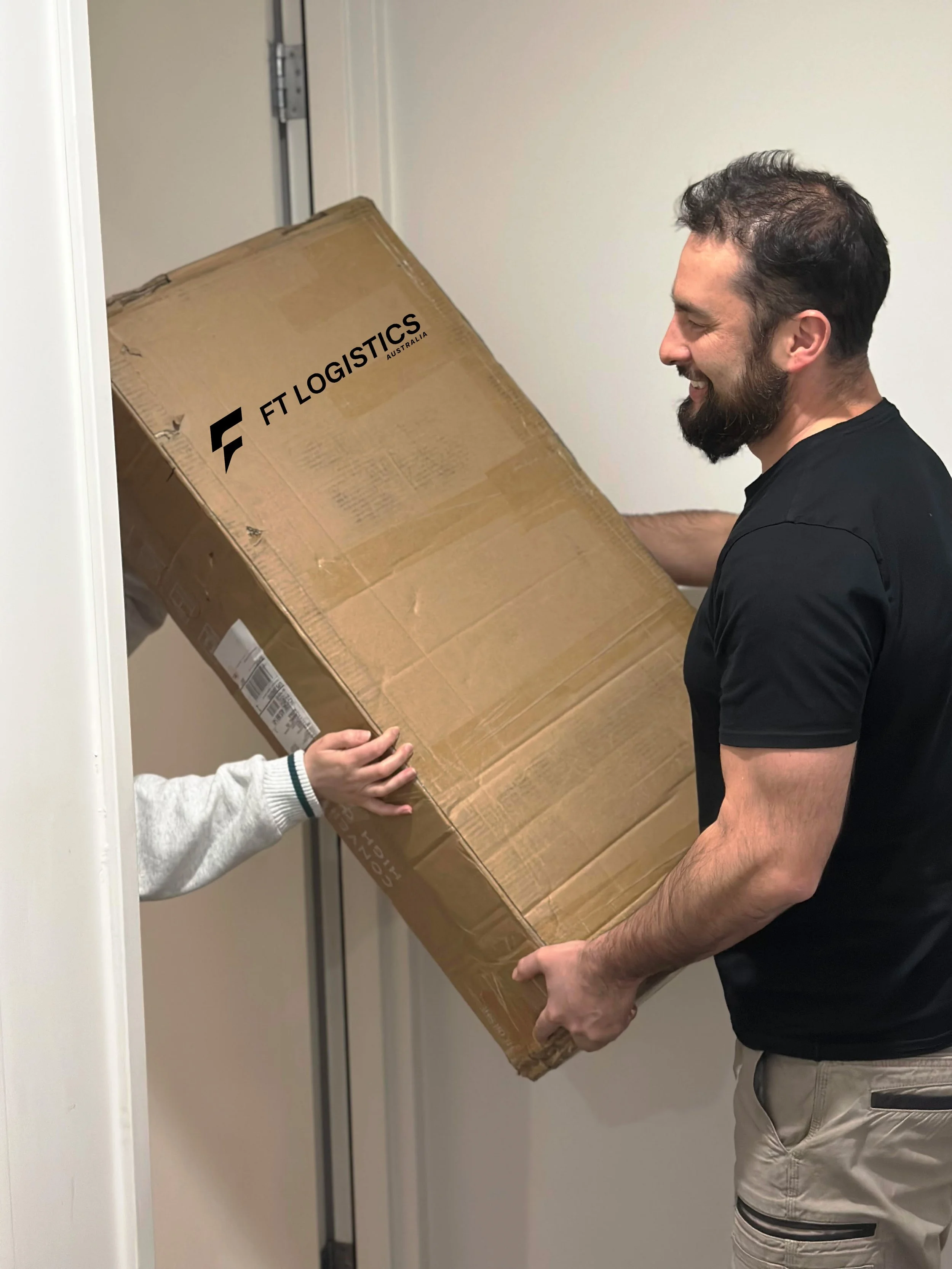 A man with a beard and black shirt smiling as he holds a large cardboard box labeled 'FTLogistics Australia' while a person's hand helps support the box in an indoor setting.