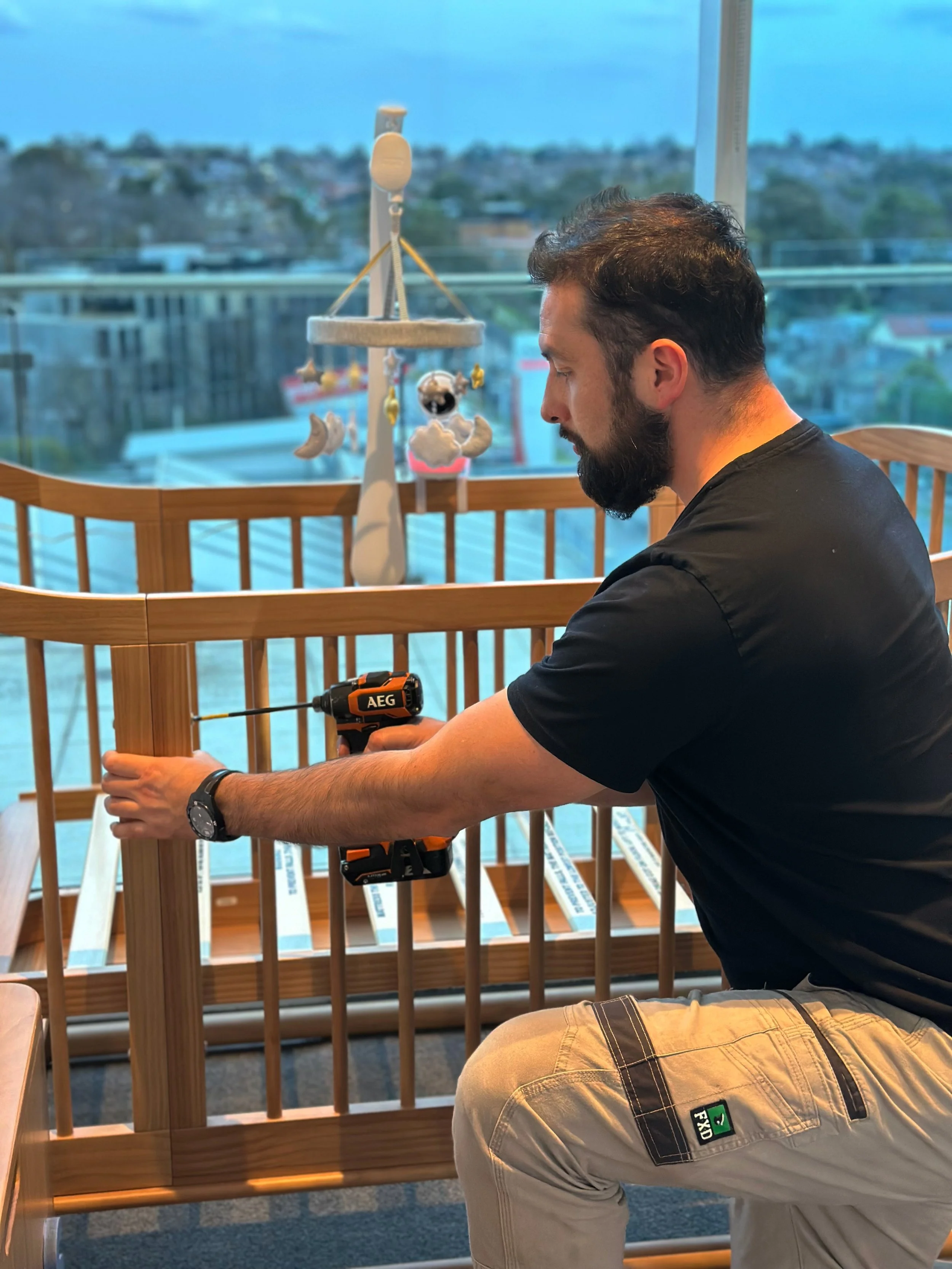 Man assembling a wooden crib with a cordless drill in a bright room with large windows and a city view in the background.