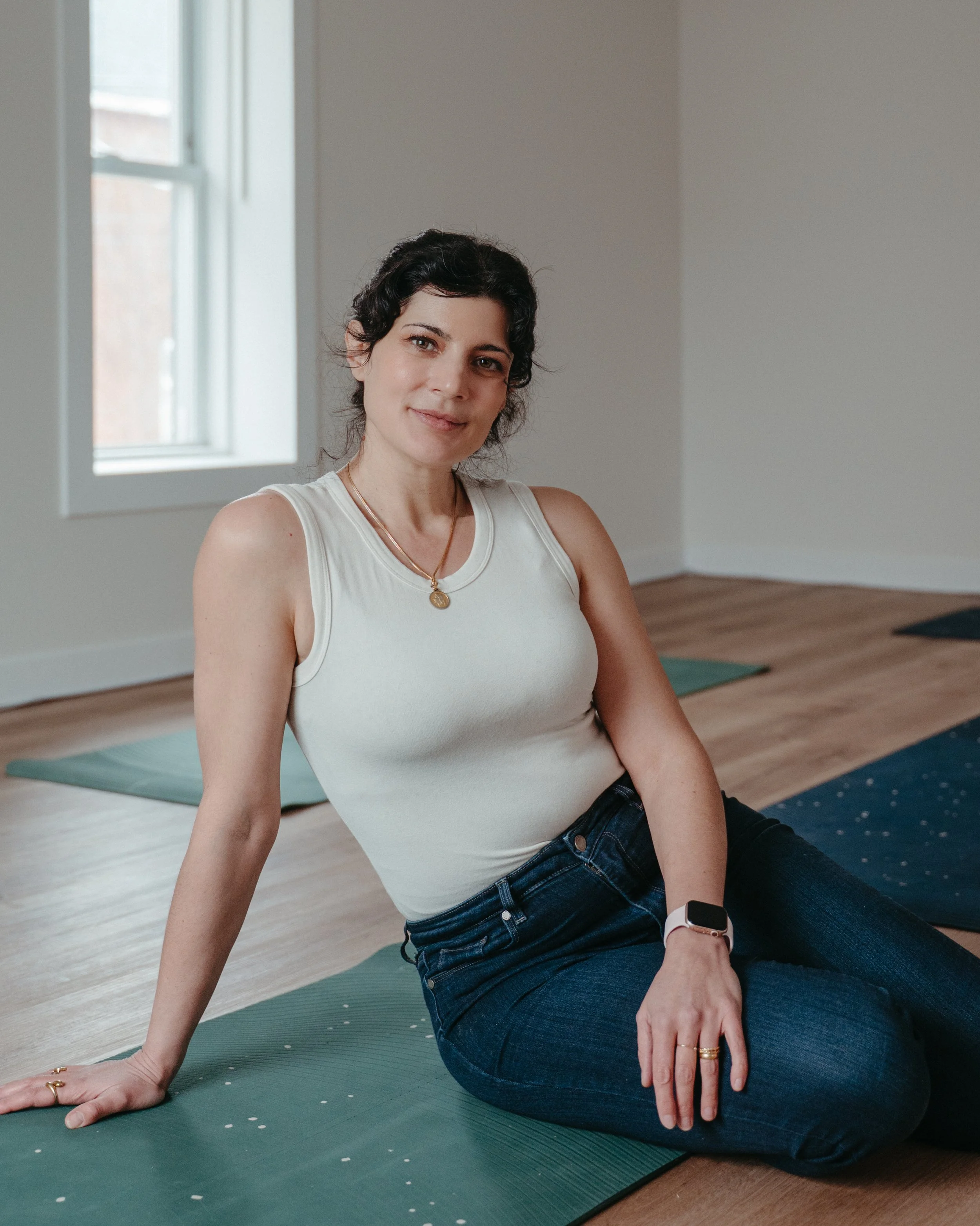 Woman sitting on yoga mat in a room with wooden floor and a window, wearing a white tank top, jeans, and a smartwatch.