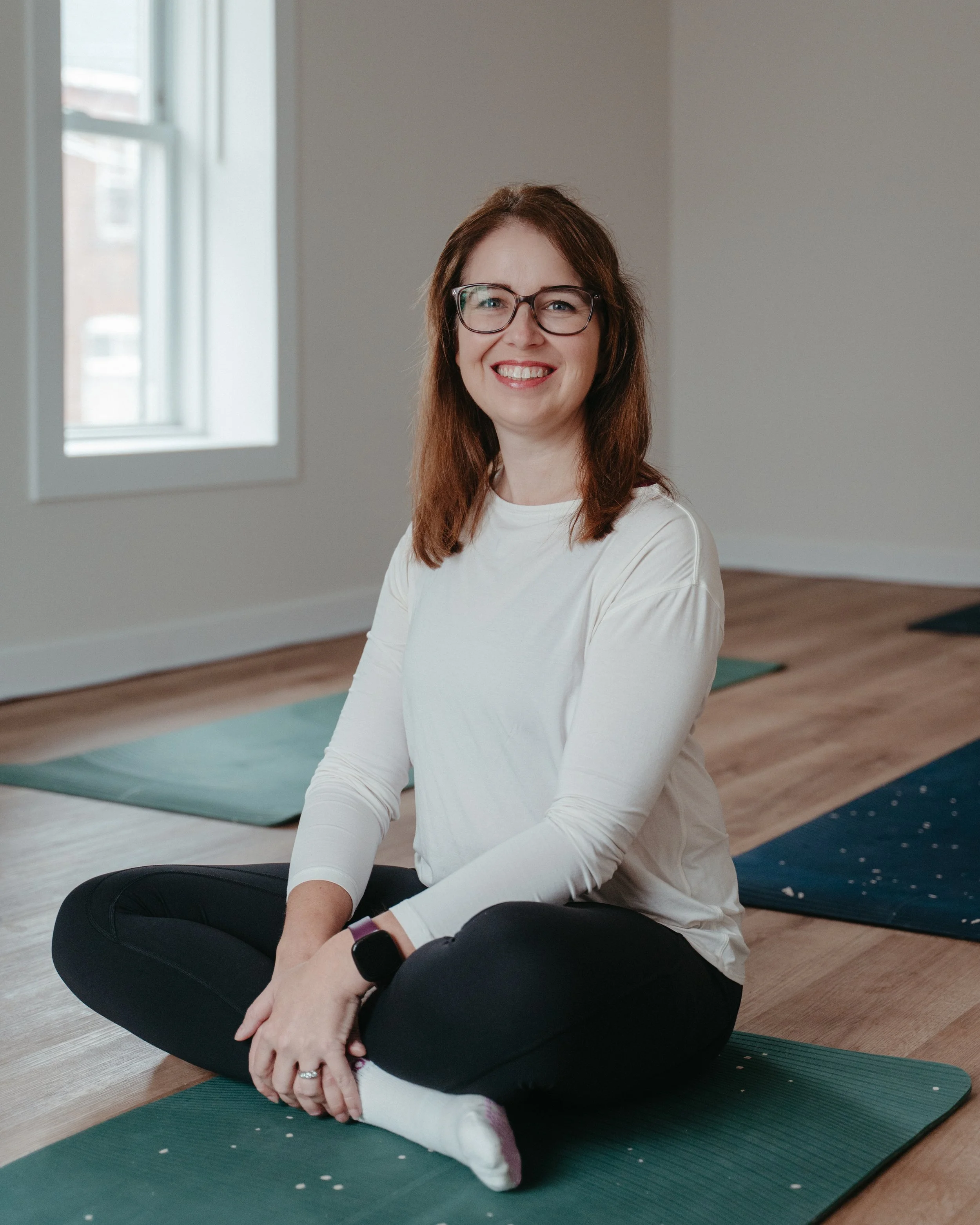 A woman with glasses sitting cross-legged on a yoga mat in a bright room, smiling at the camera.