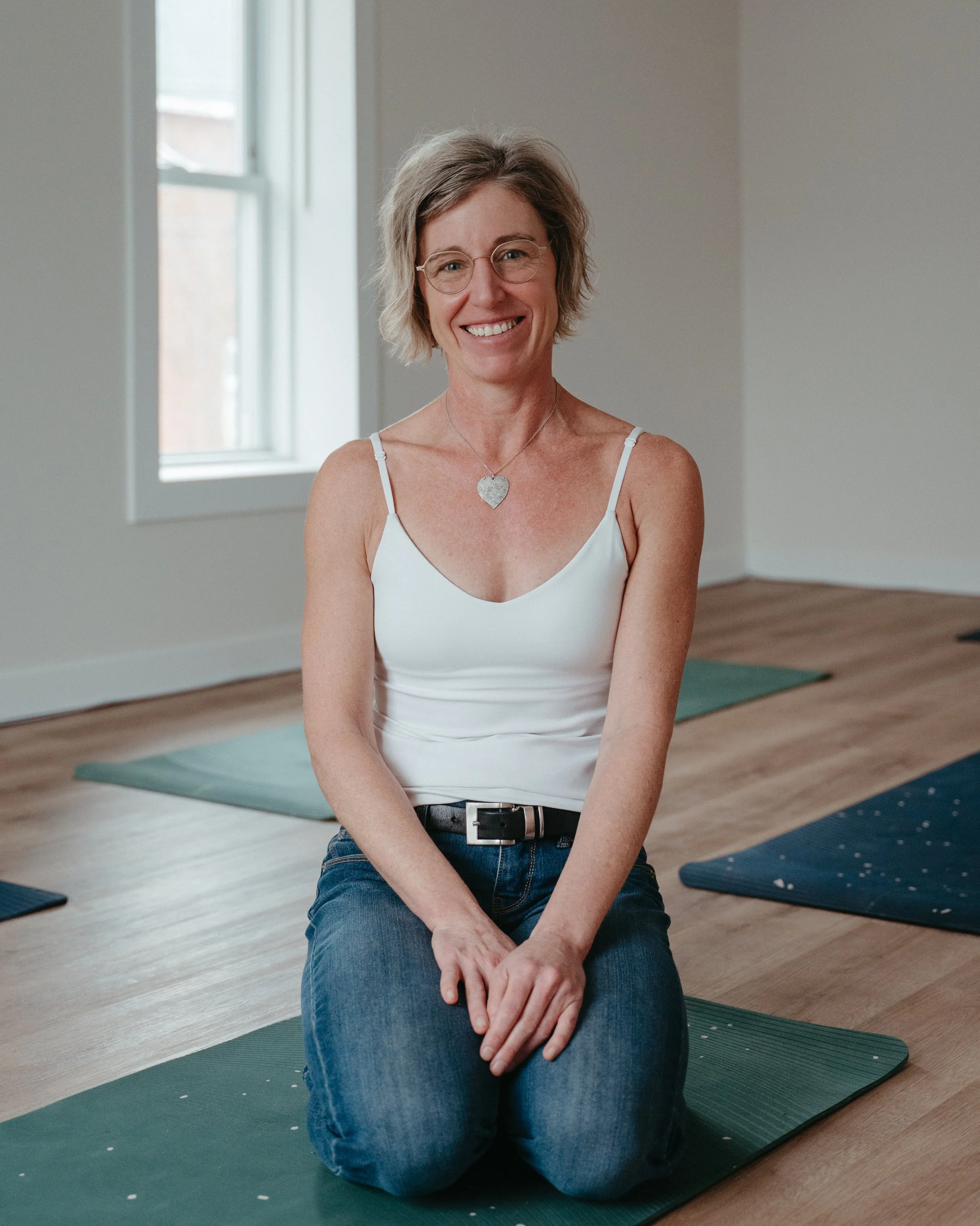 A woman sitting on a yoga mat in a bright room with large windows, smiling at the camera.