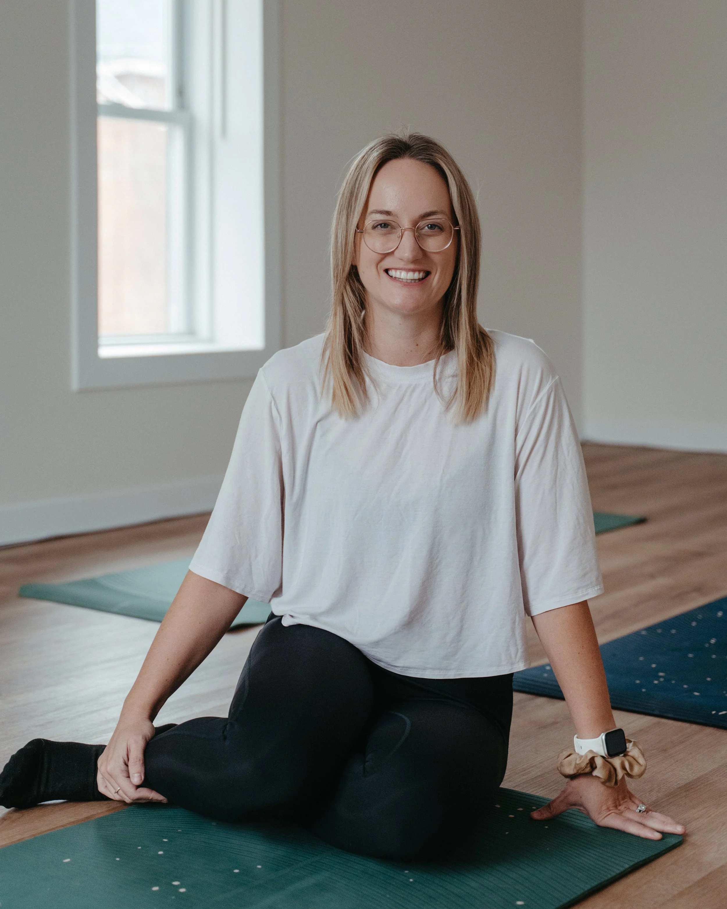 A smiling woman sitting on a yoga mat in a bright indoor space, preparing for yoga or exercise.