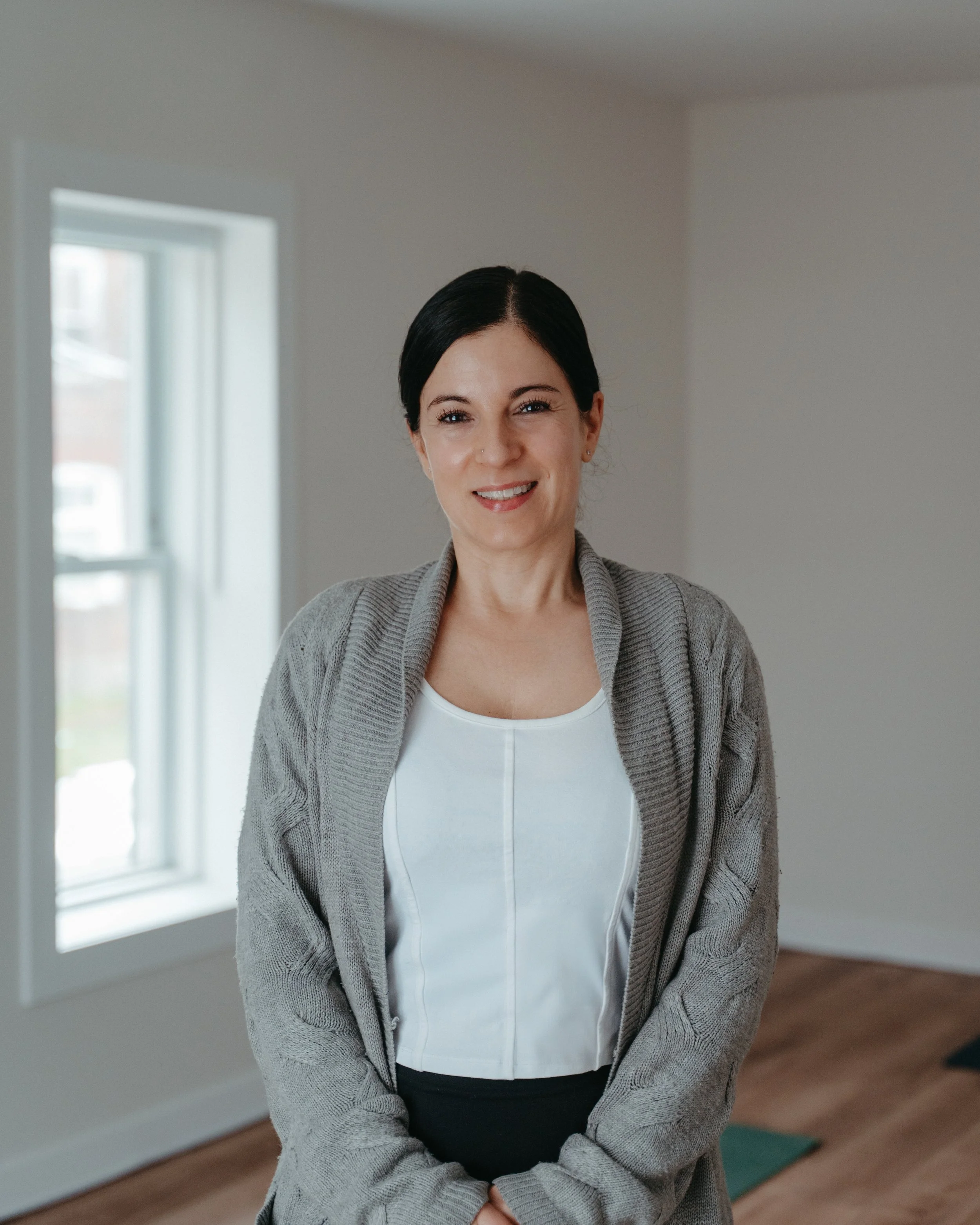 A woman with black hair tied back, smiling, wearing a light gray cardigan over a white top, standing inside a room with a window and a wooden floor.
