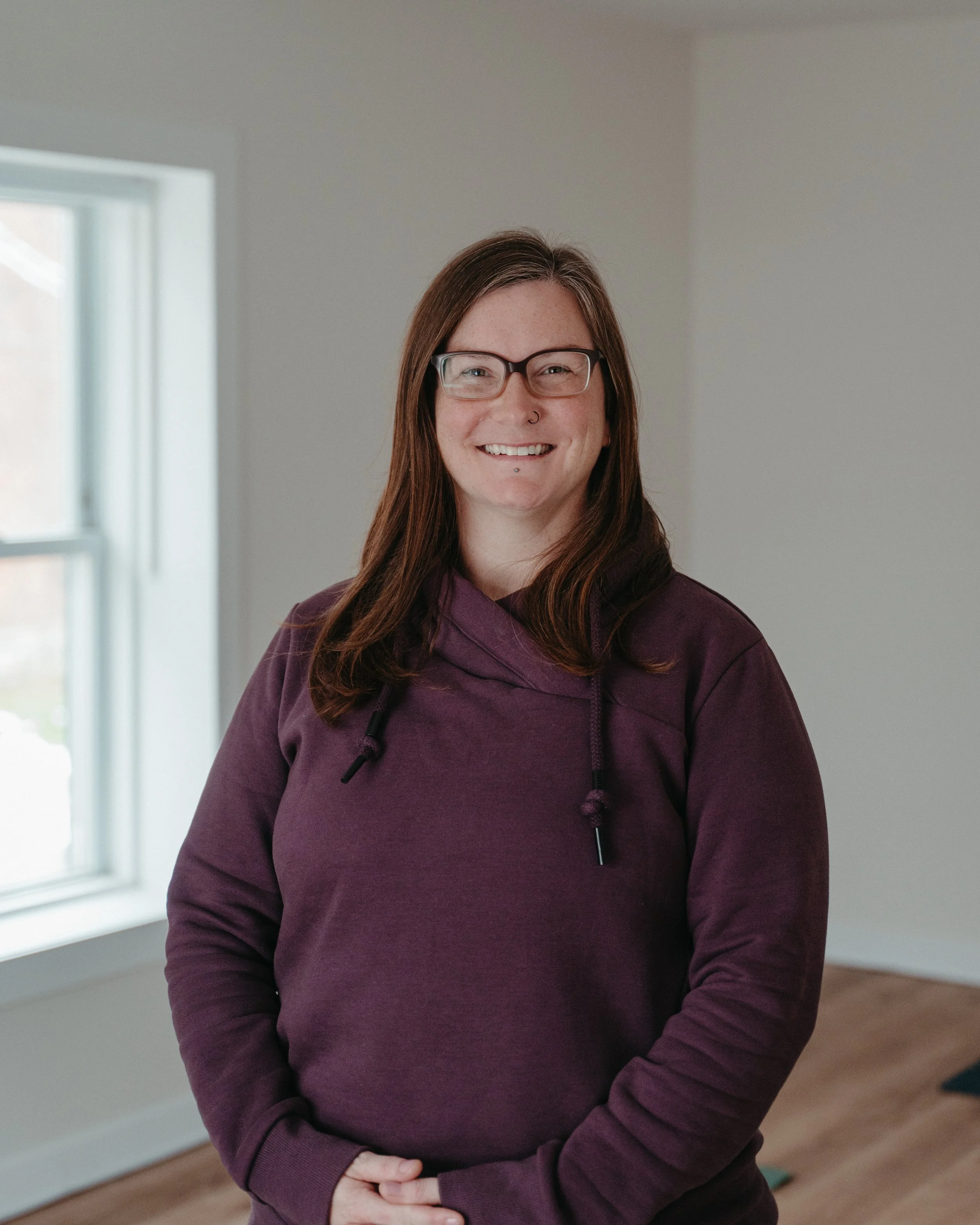 A woman with long brown hair, glasses, and a nose piercing, smiling while wearing a purple hoodie, standing indoors near a window.