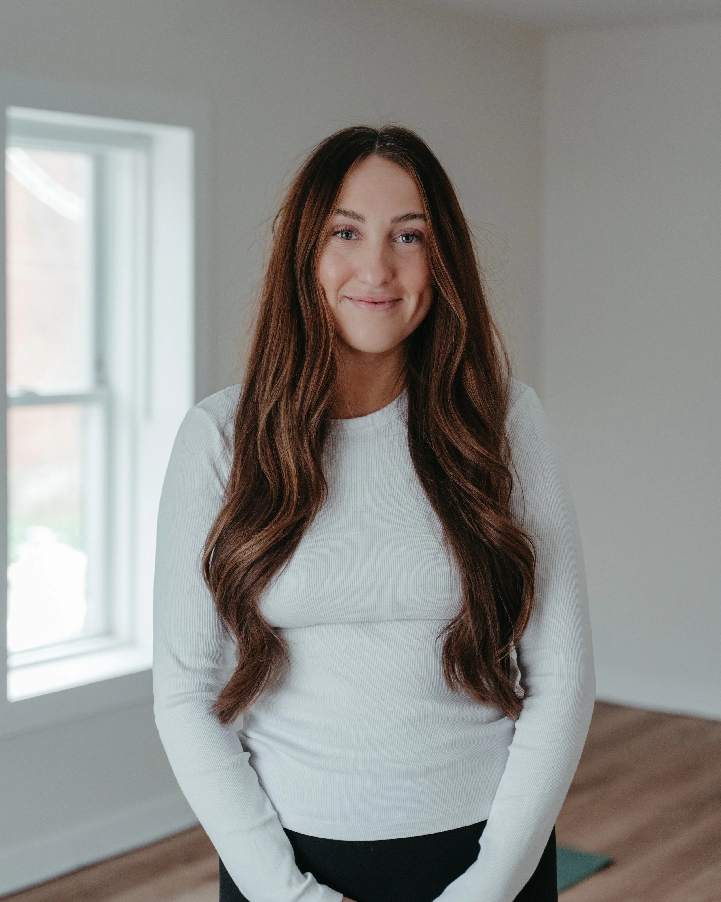 Young woman with long brown hair smiling and standing indoors in front of a window.