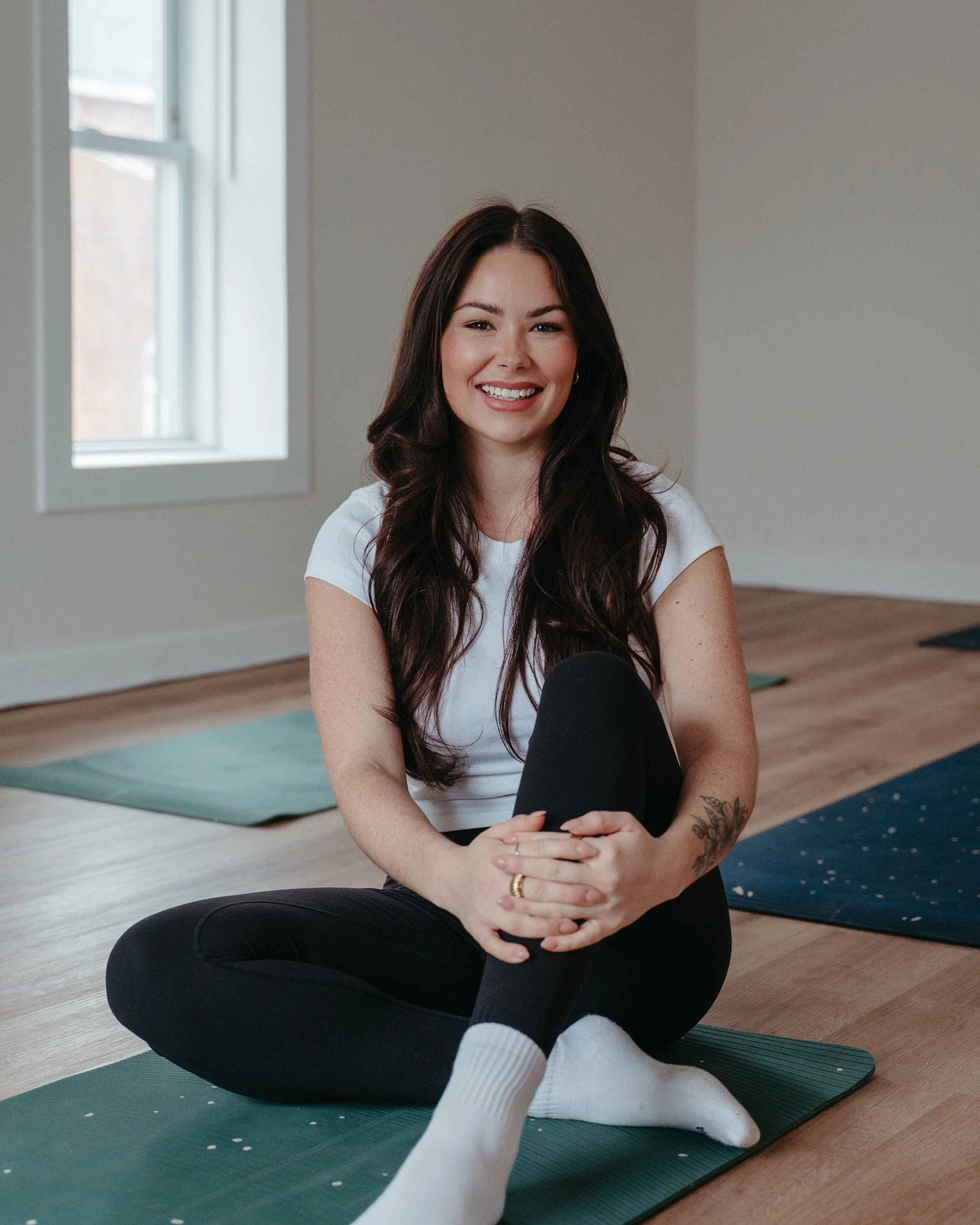 A woman smiling and sitting on a yoga mat in a room with hardwood floors and a window, wearing a white t-shirt, black pants, and white socks.
