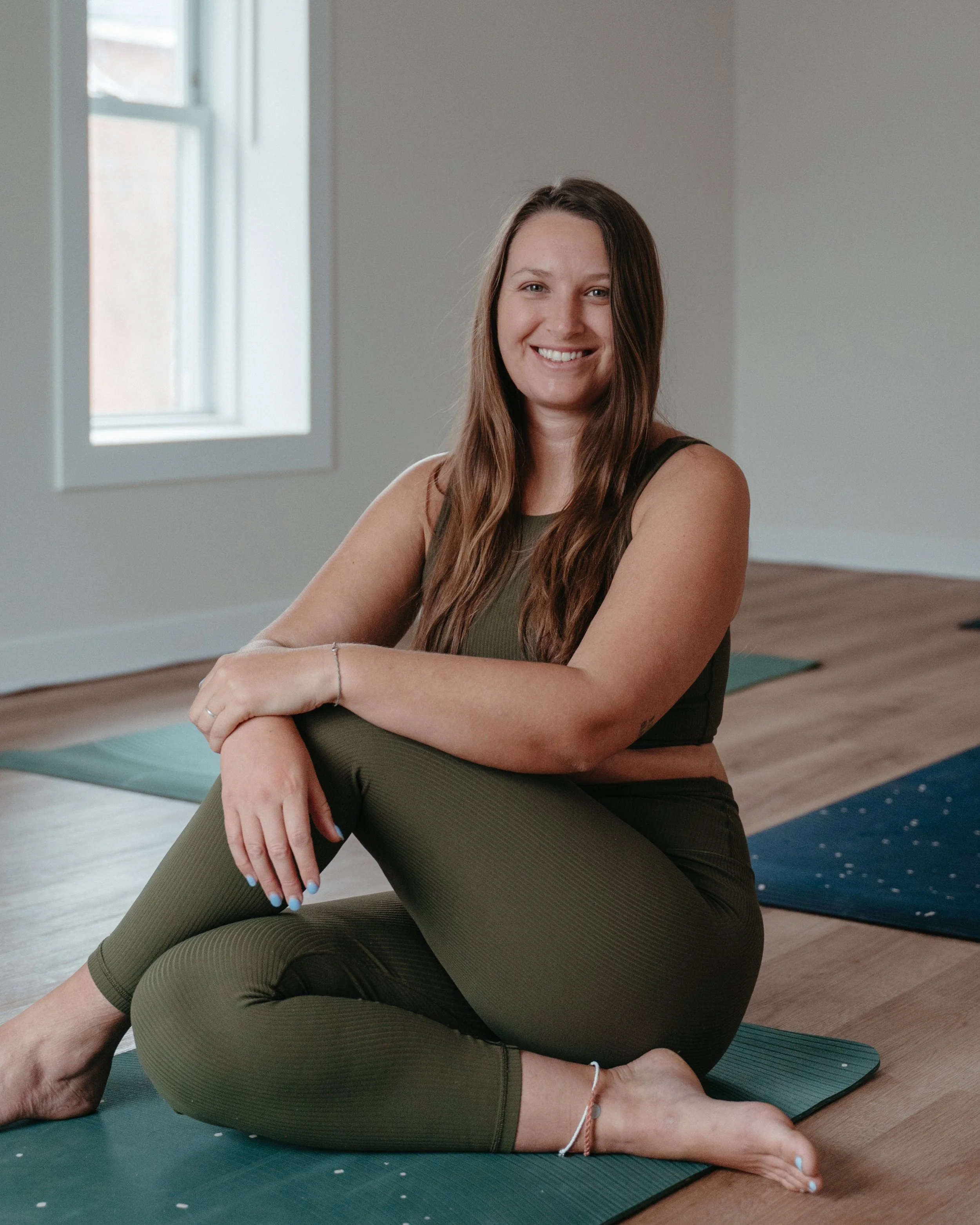 A woman sitting on a yoga mat in a room with light-colored walls and wooden flooring, smiling at the camera.