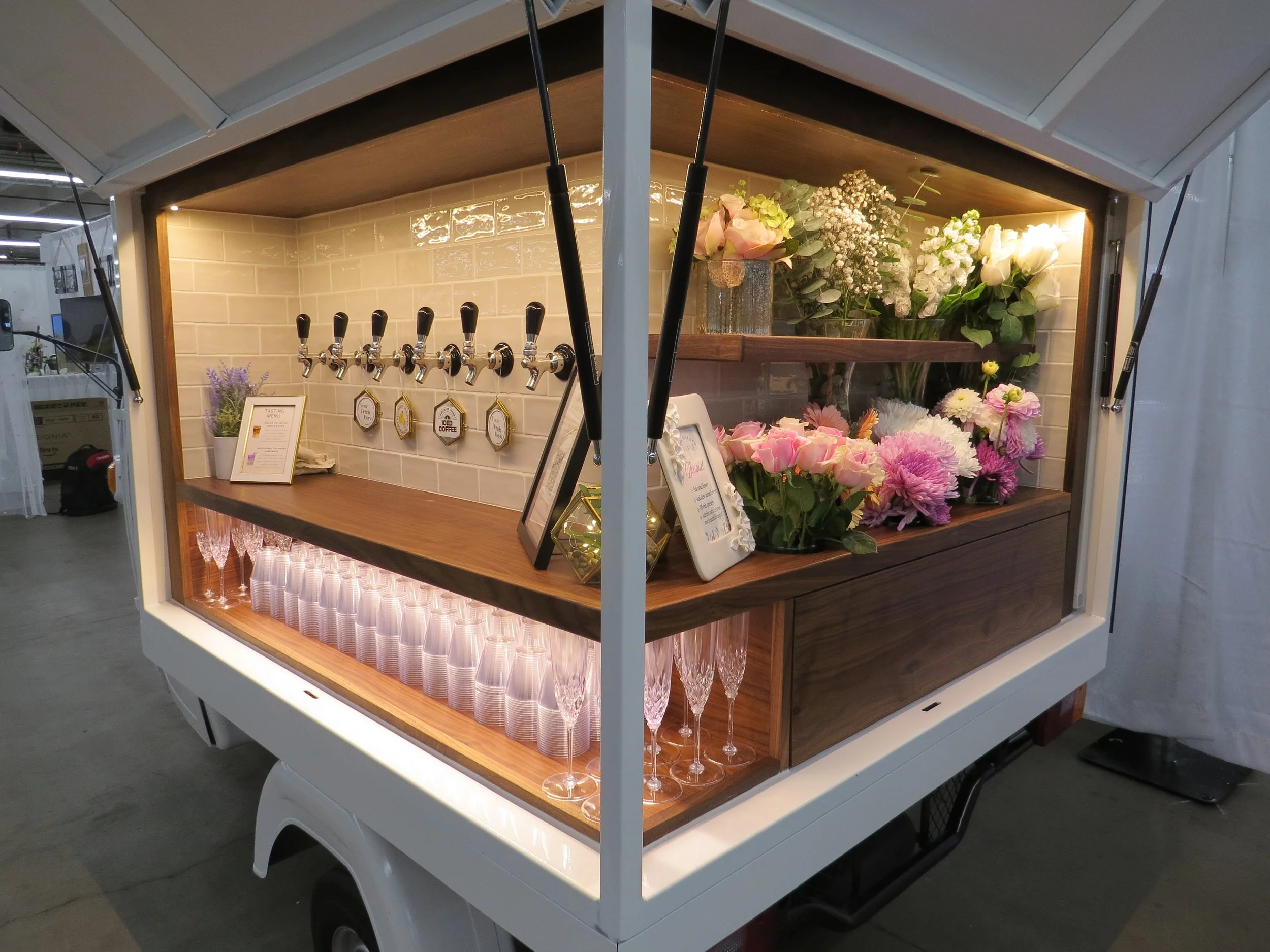 Two clear plastic cups, one filled with a golden-colored beverage and ice cubes, placed on a wooden surface next to a beige fabric, with a background of pink and white flowers and green foliage.