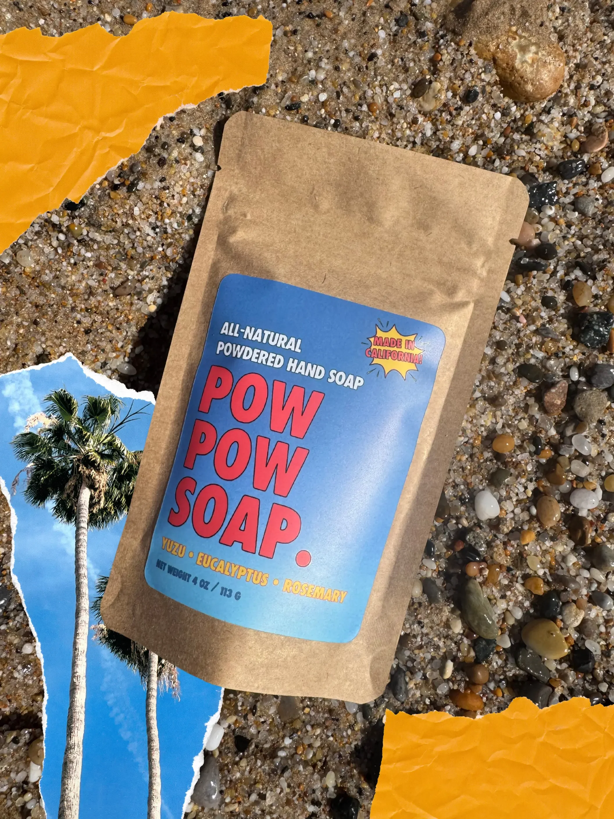 A package of Pow Pow Soap, all-natural powdered hand soap, with ingredients including yuzu, eucalyptus, and rosemary, placed on a sandy beach with small rocks, along with torn pieces of yellow paper and a torn piece of paper with a palm tree against a blue sky.