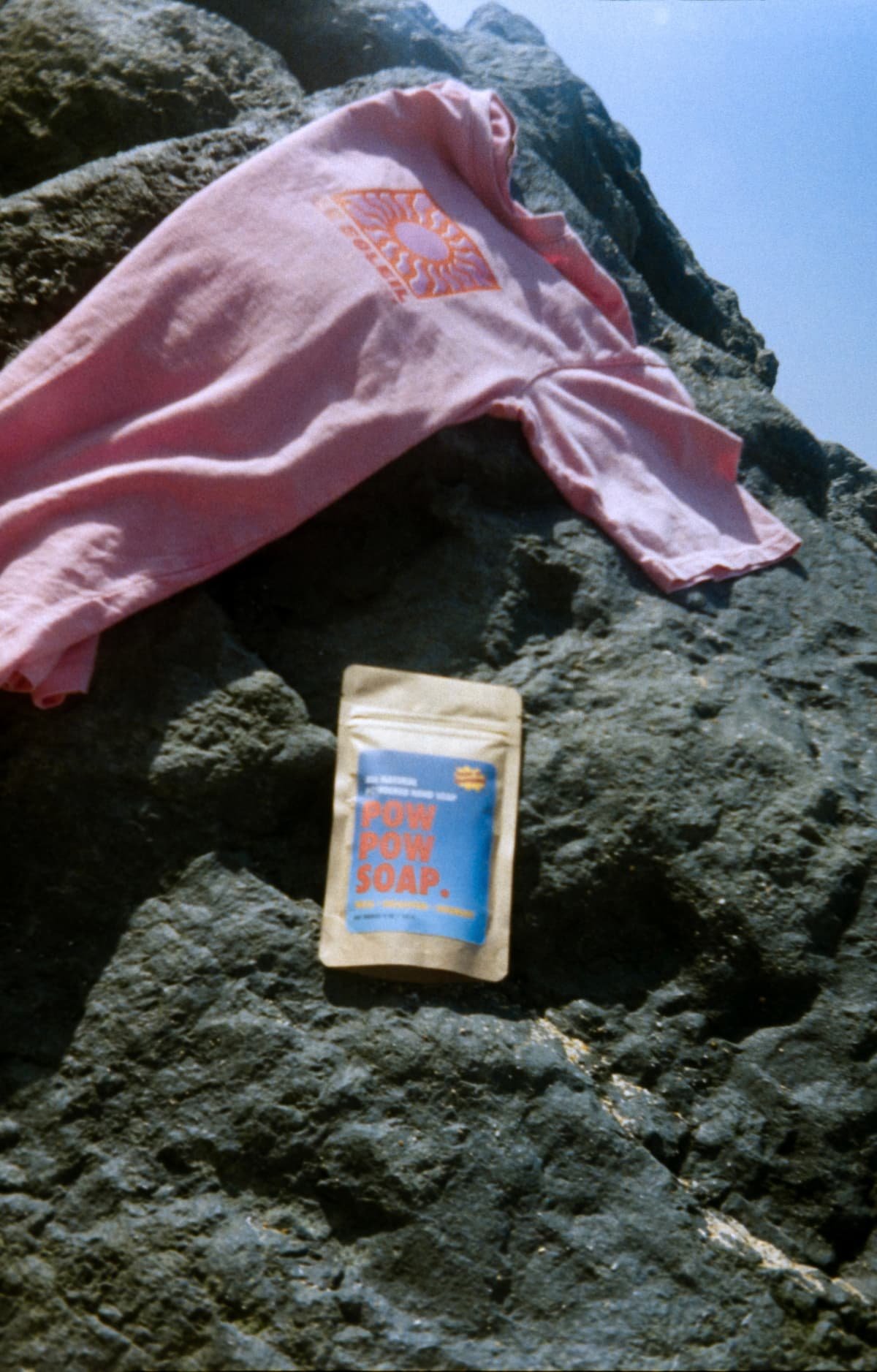Pink towel and a package of POW POW SOAP placed on a rocky surface outdoors.