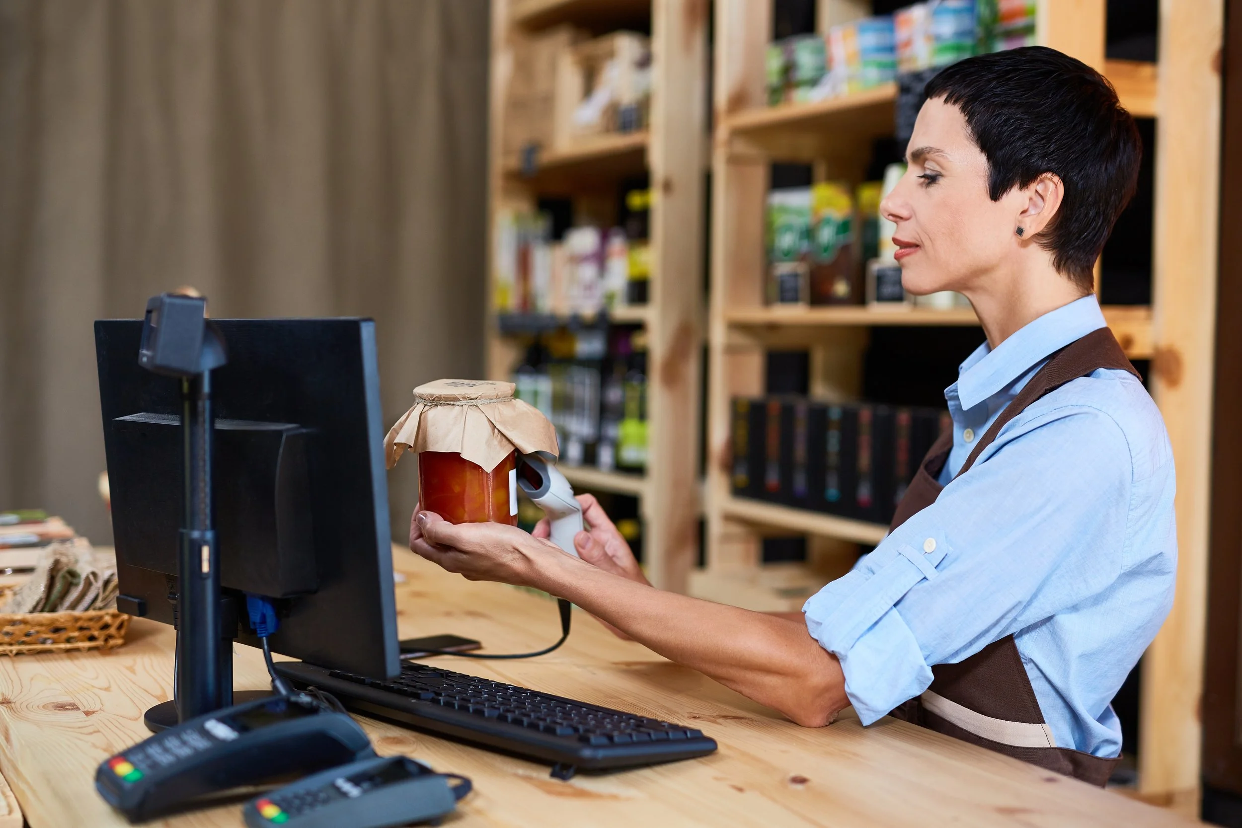 A woman works behind a wooden counter with a computer, bar code scanners, and payment terminal, holding a jar of jam in a store with shelves of products in the background.