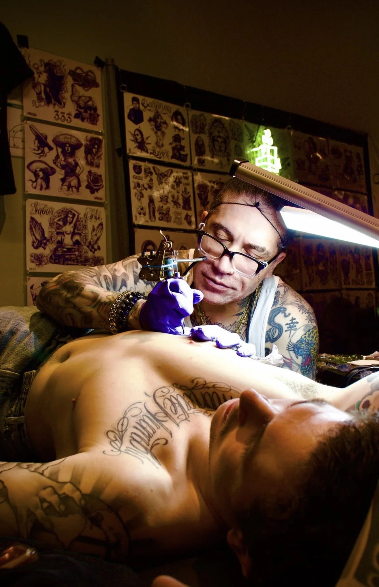 A tattoo artist is tattooing a person who is lying on a table. The artist is wearing glasses, gloves, and has tattoos on his arms and neck. The tattooed person has tattoos on their chest and arm, and their head is turned to the side. The background features wall art with various tattoo designs and a bright green neon sign.