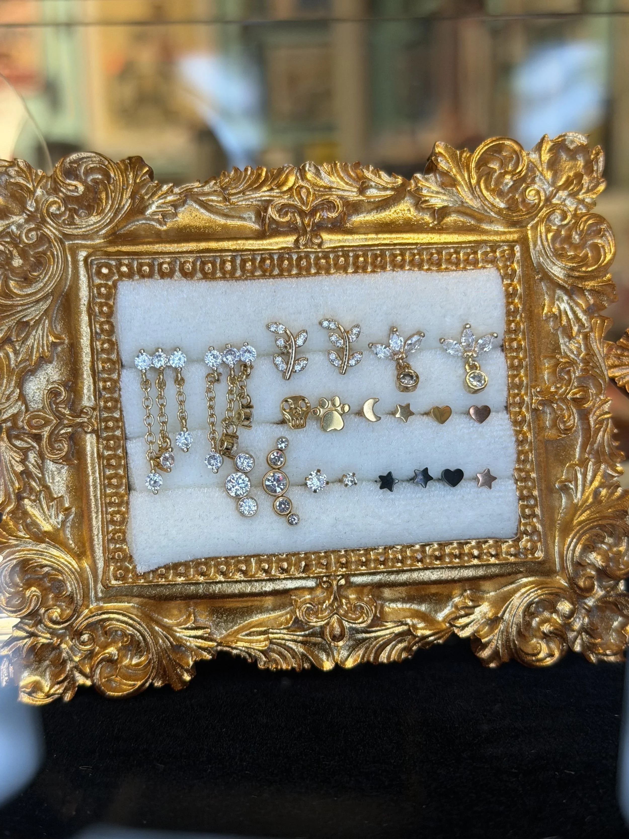 A collection of small jewelry pieces, including earrings with various designs, displayed on a white cushioned background inside an ornate gold frame.