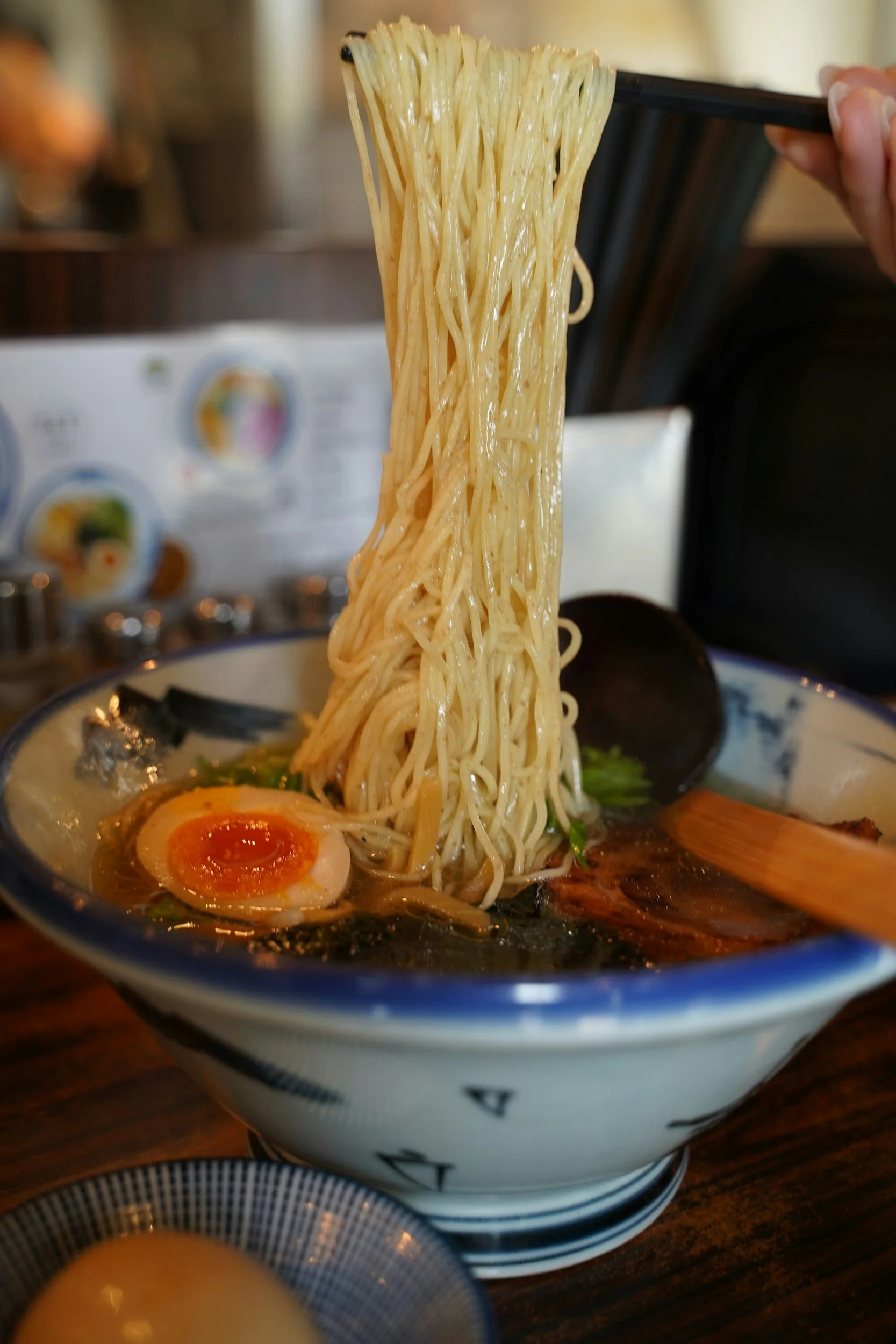 A bowl of ramen with noodles being lifted by chopsticks, topped with a soft-boiled egg, slices of meat, and green vegetables.