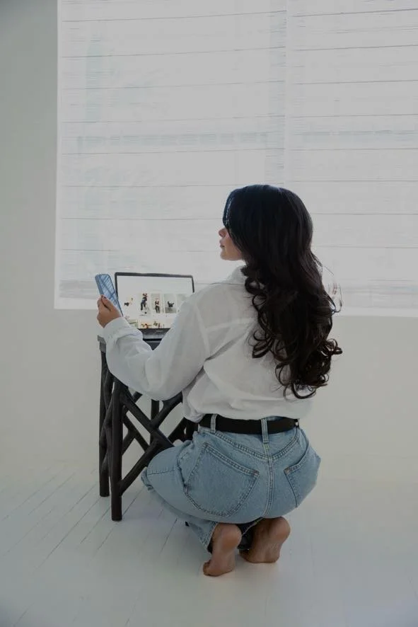A woman with long dark hair in a white shirt and light jeans, kneeling on the floor, looking at her phone and a laptop, in front of a large window with blinds. blockstyle-border