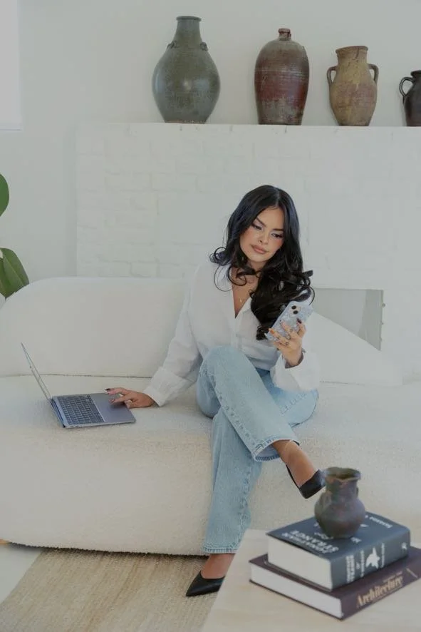Woman sitting on a white sofa, holding a smartphone, with a laptop beside her, in a room with decorative vases on a shelf above. blockstyle-border