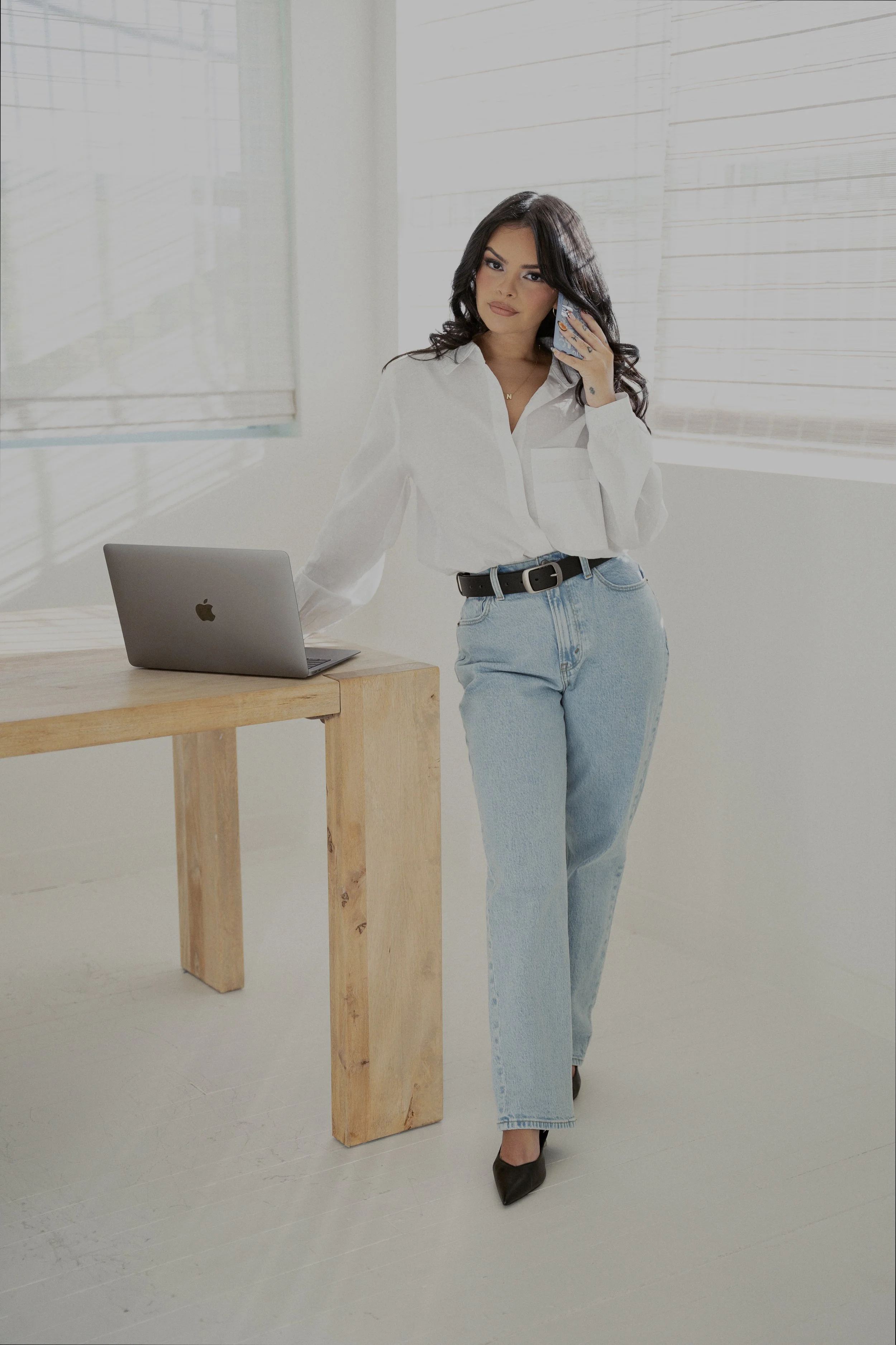 A woman with long dark hair, wearing a white blouse, light blue jeans, and black high heels, stands by a wooden table, talking on a cellphone inside a bright room with white walls and blinds. blockstyle-border