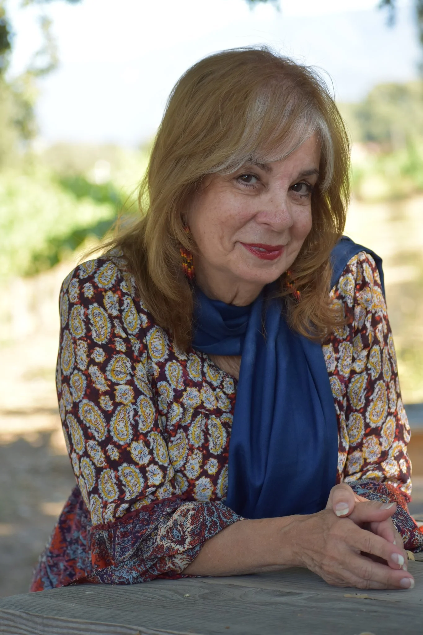 Close-up of an elderly woman with blonde hair, wearing a patterned blouse with a blue scarf, sitting outdoors at a wooden table with her hands clasped.