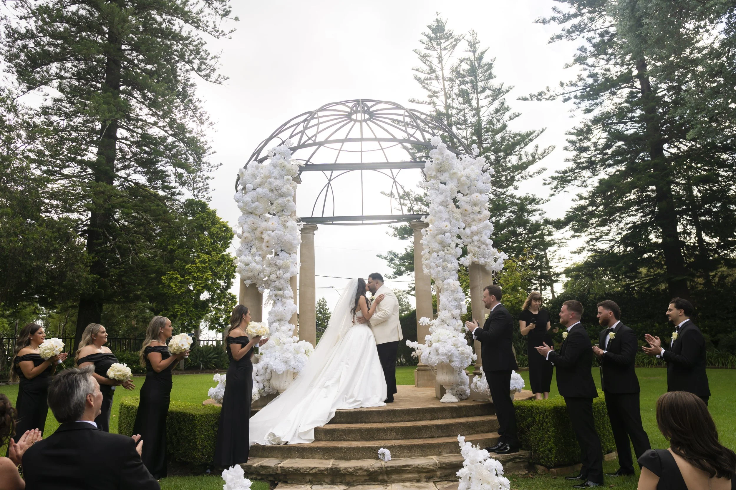 A wedding ceremony outdoors with a bride and groom kissing under a decorative arch surrounded by wedding party and guests, with tall trees in the background.