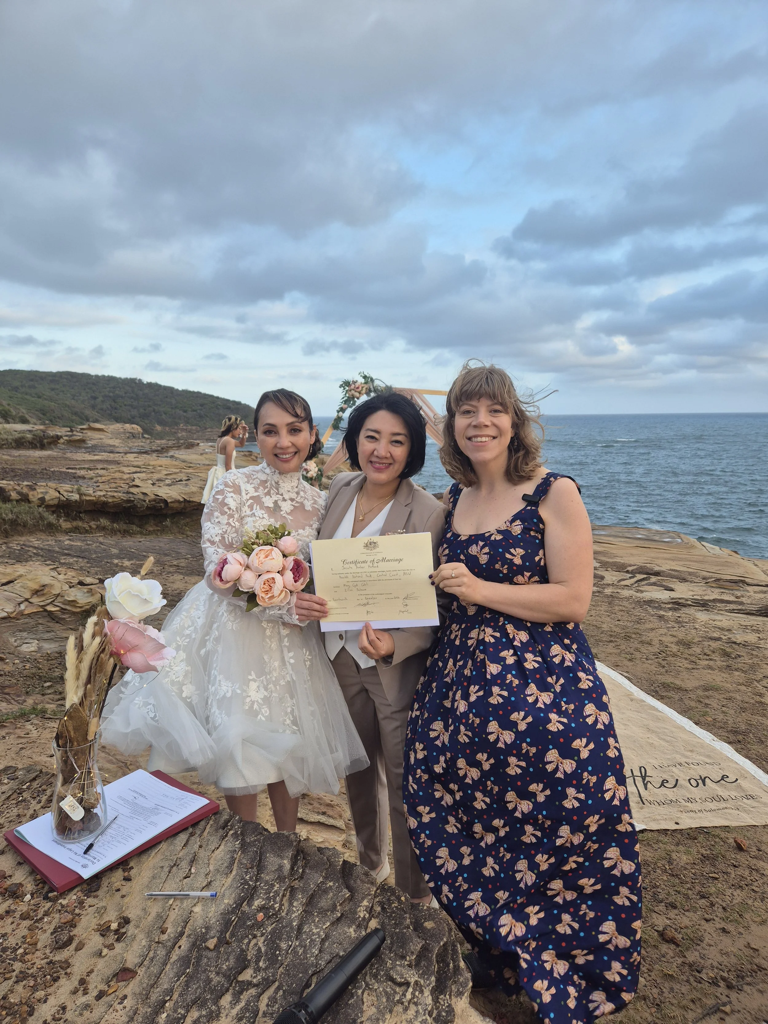 Three women standing outdoors on a rocky coastline, one in a wedding dress holding a bouquet, holding a marriage certificate, and the other two women smiling. A woman in a beige suit and a woman in a floral dress. In the background, a woman in a whit