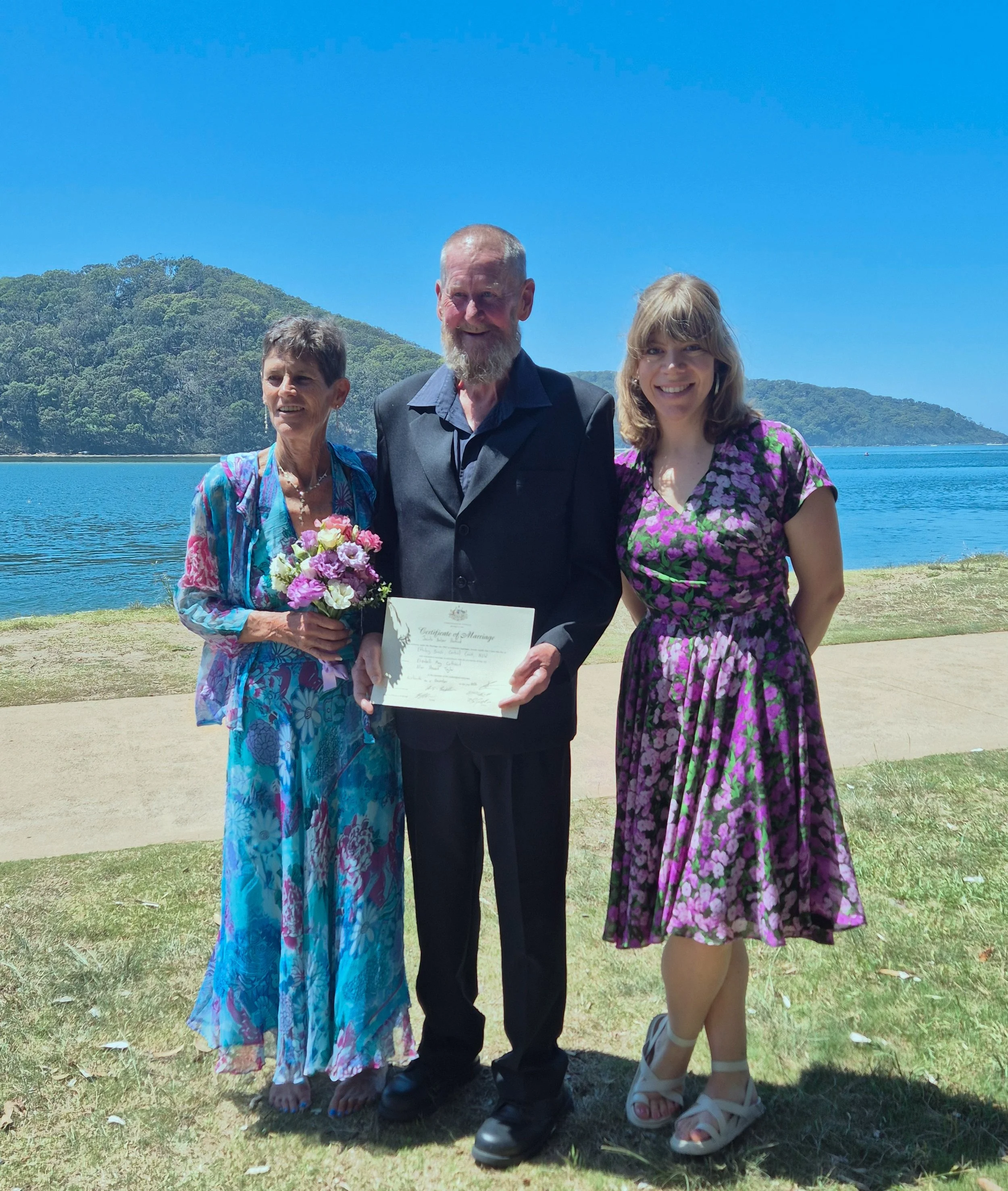 A family celebrating a wedding outdoors near a lake with a mountain in the background. The woman on the left is holding a bouquet of flowers, and the man in the middle is holding a wedding certificate. The woman on the right is smiling, wearing a pur