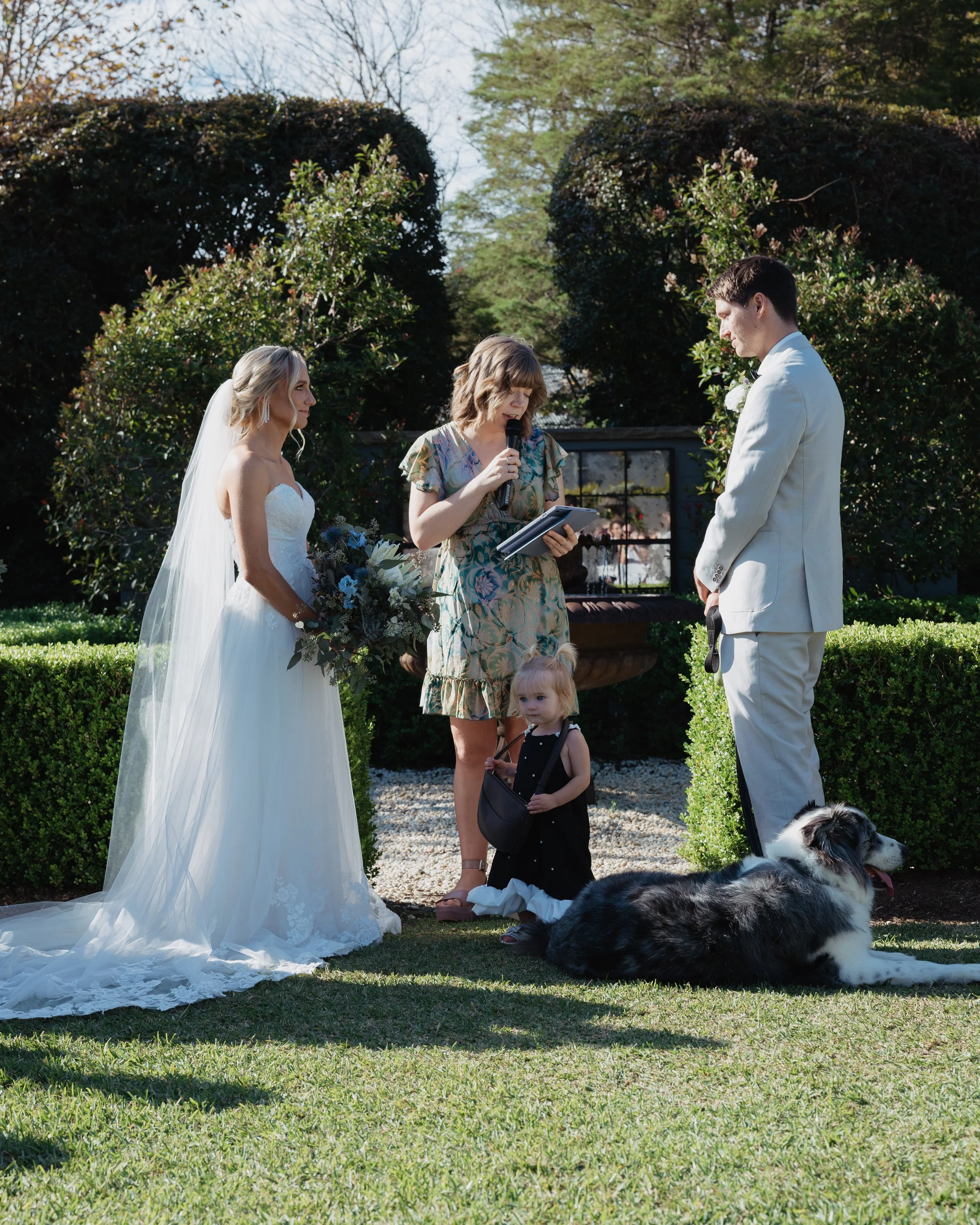 Bride and groom at outdoor wedding ceremony with officiant, a young girl, a small dog, and lush garden background.