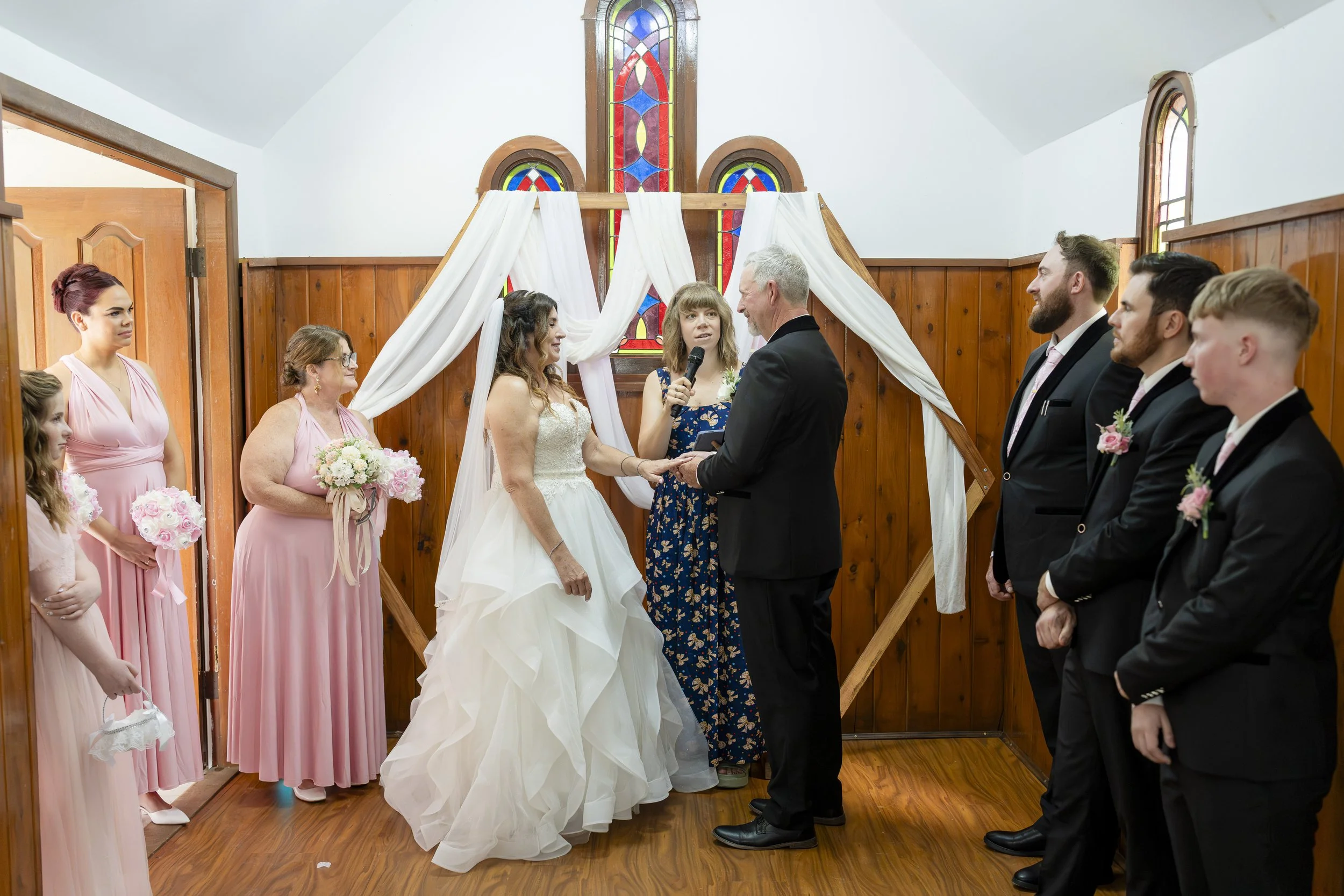 A wedding ceremony taking place indoors with a bride and groom exchanging vows in front of a priest, surrounded by bridesmaids and groomsmen. The bride is in a white wedding gown, and the groom is in a black suit. Bridesmaids are in pink dresses, and