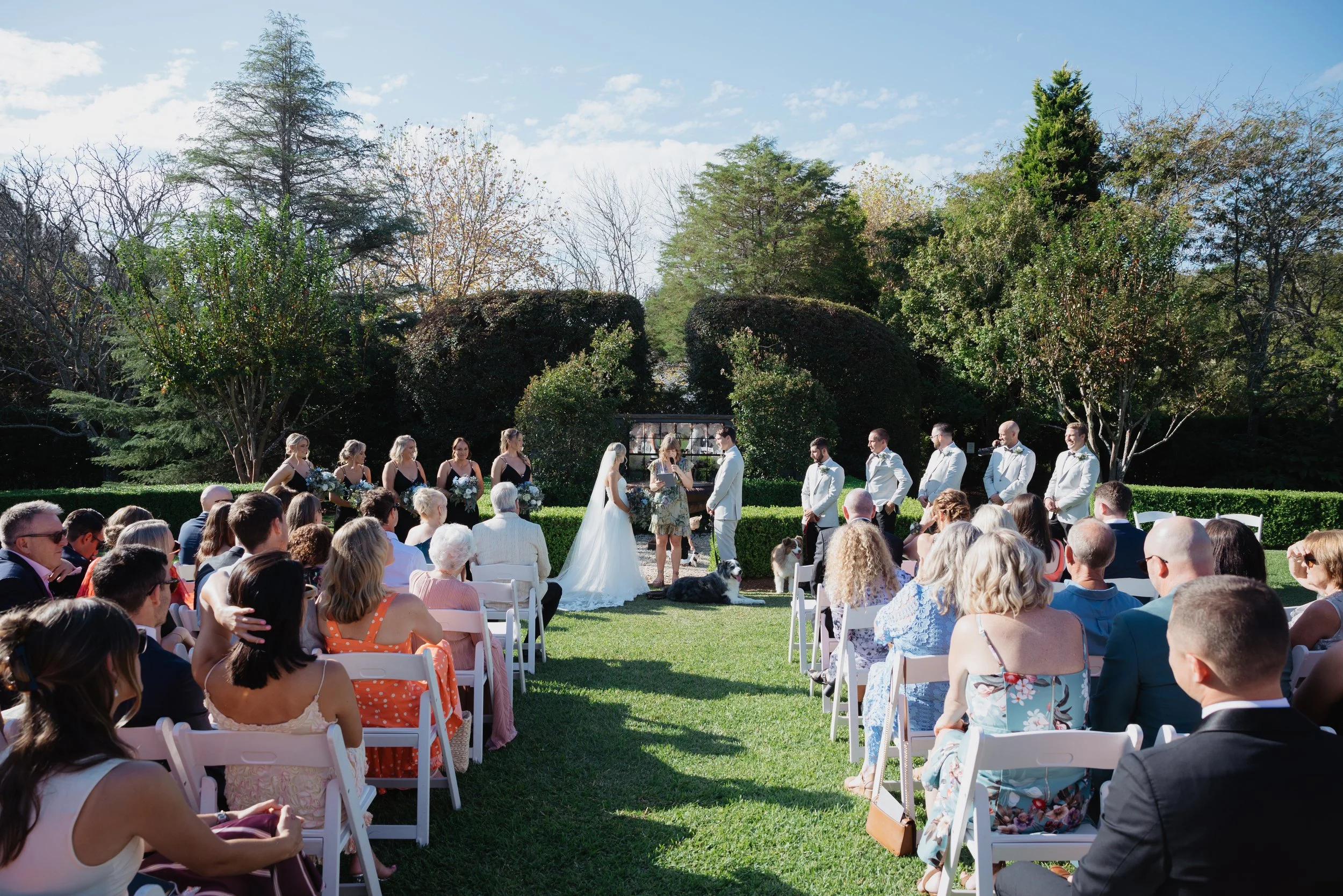Outdoor wedding ceremony with bride, groom, officiant, bridesmaids, groomsmen, and seated guests on a sunny day in a garden setting.