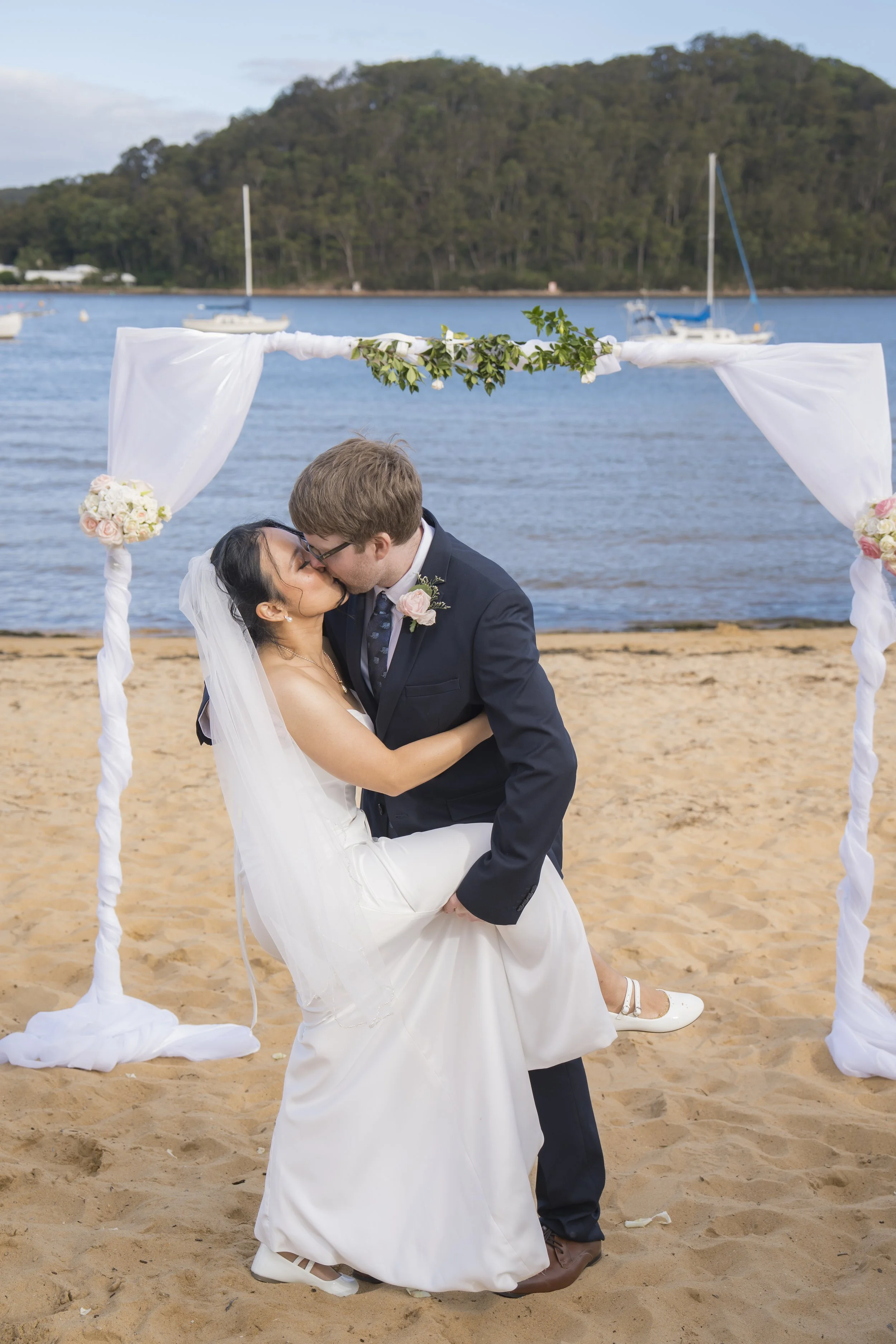 A newlywed couple kissing under a wedding arch on a sandy beach with boats on calm water and a forested hill in the background.