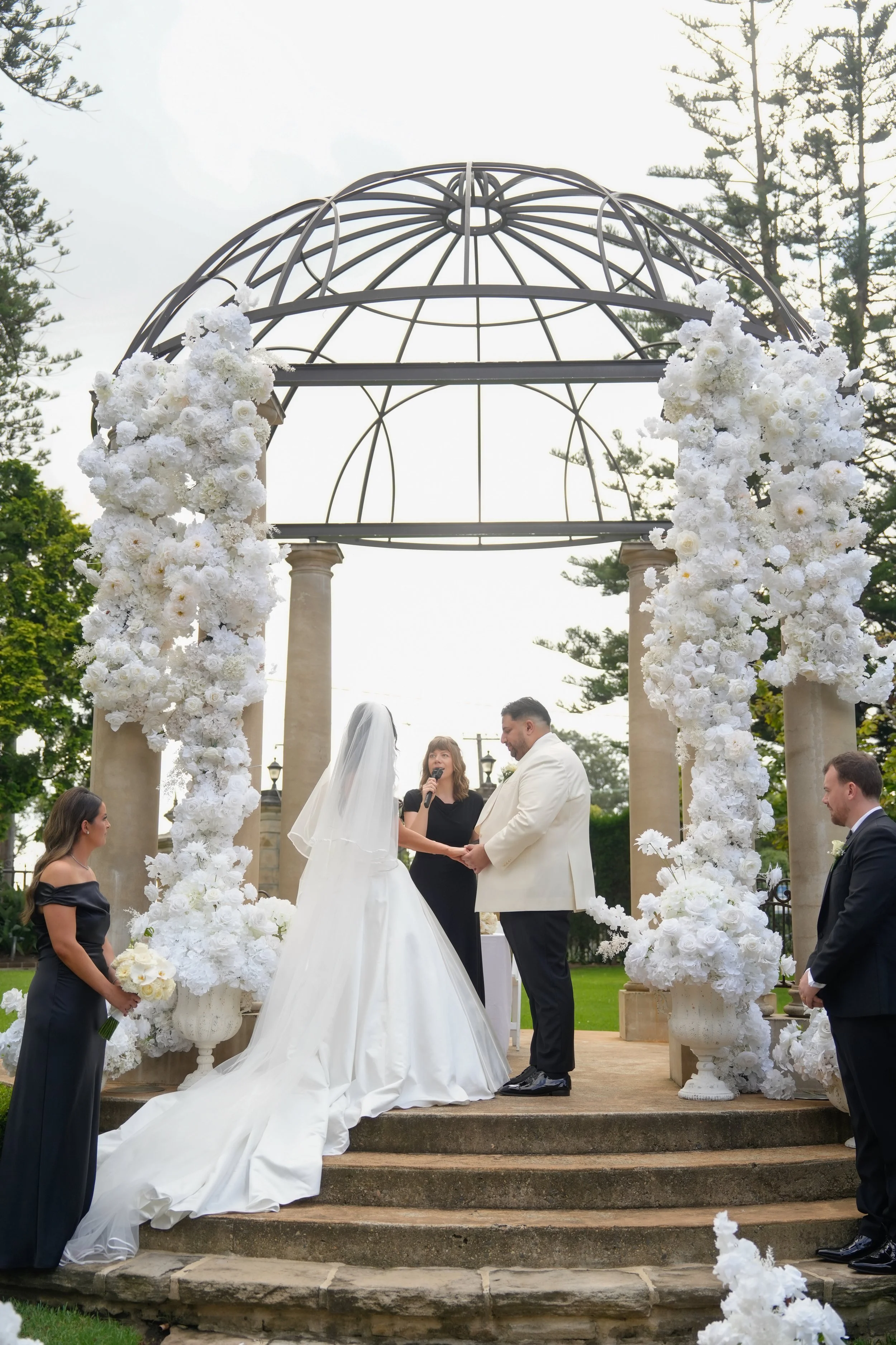 A wedding ceremony taking place outdoors under a decorative wrought iron archway adorned with white flowers. The bride and groom are holding hands and exchanging vows, surrounded by attendants.