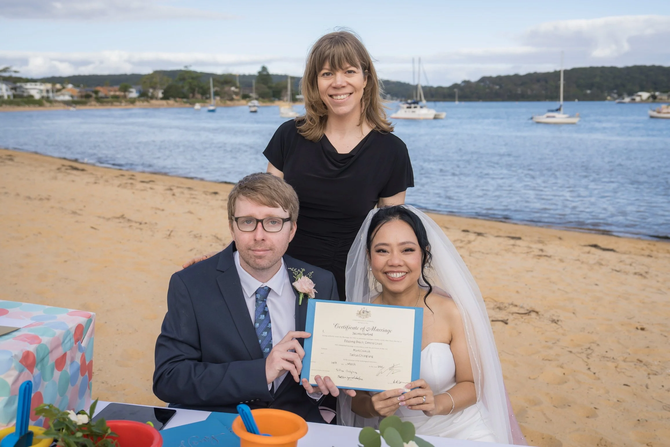 A couple at a beach wedding holding a marriage certificate, with a woman in a white wedding dress and veil and a man in a dark suit, alongside two women, one in black with brown hair and the other with glasses and light brown hair. In the background,