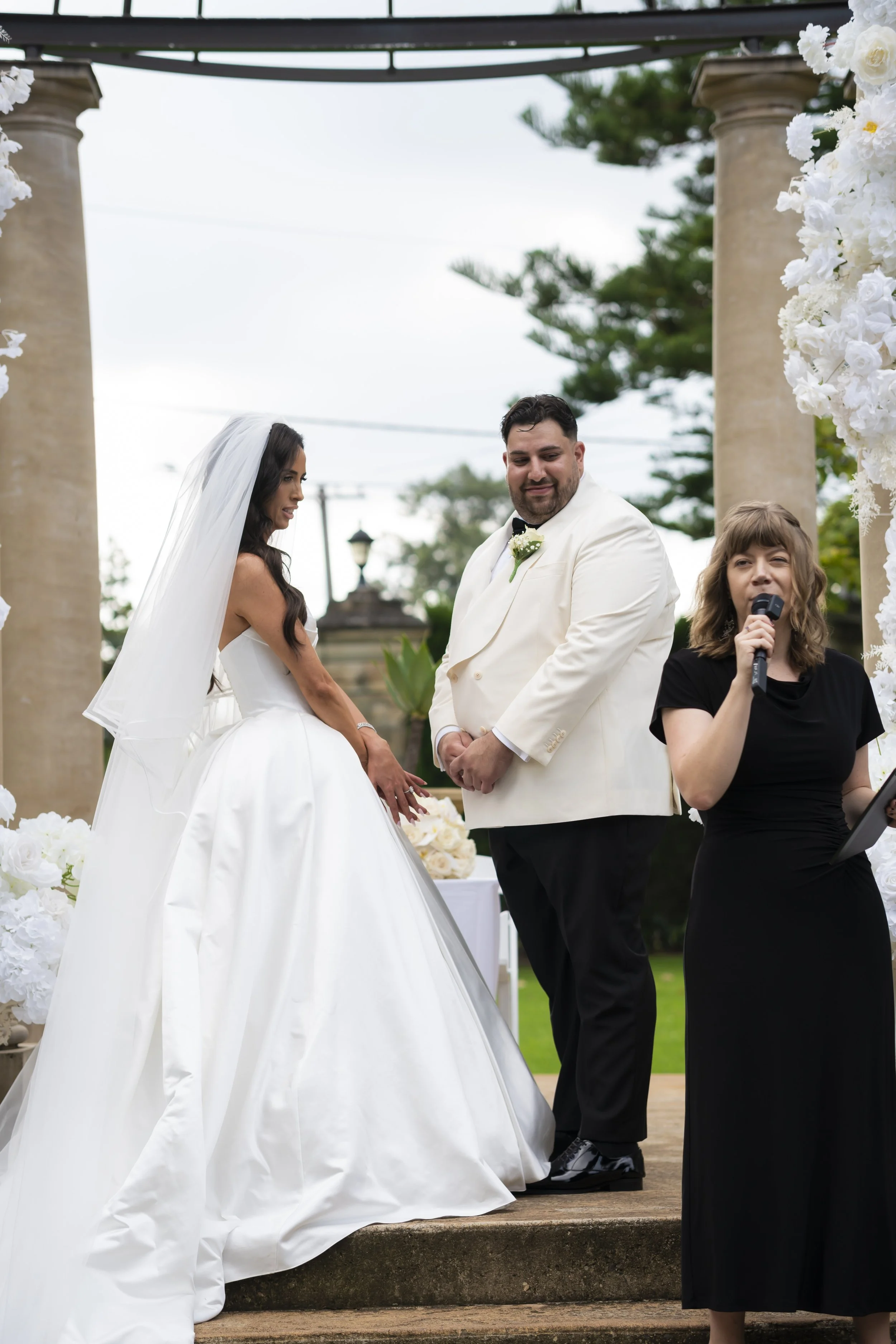 A bride and groom exchanging vows during an outdoor wedding ceremony, with a woman officiant holding a microphone.