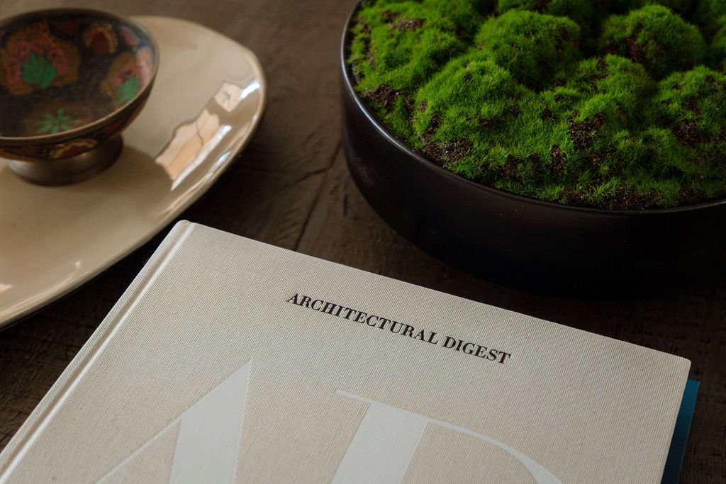 A hardcover book titled 'Architectural Digest' placed on a wooden table, next to a black bowl filled with green moss and a white plate with a decorative bowl on top.